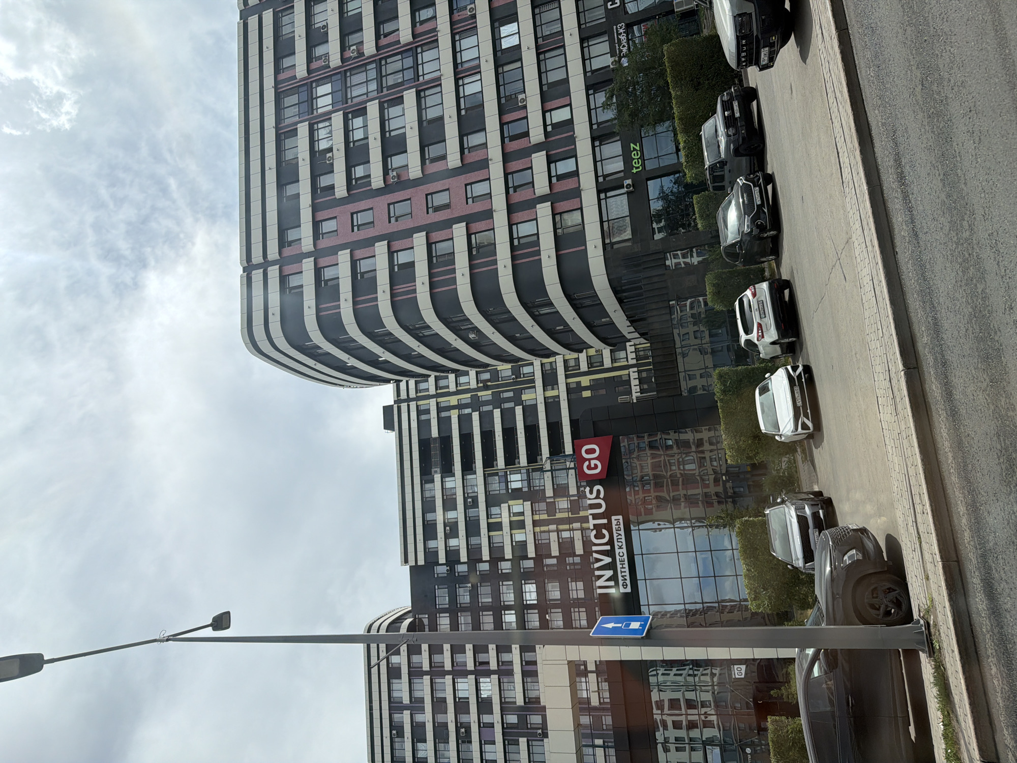 The image shows a street view in Astana, Kazakhstan, with modern multi-story residential buildings on the right and a large building with a sign that reads "INVICTUS GO ФИТНЕС КЛУБЫ" (INVICTUS GO FITNESS CLUBS) on the left. Several cars are parked in front of the buildings, including a black SUV, a white SUV, a silver SUV, and other vehicles partially visible. A street lamp is prominent in the foreground on the left. The sky is overcast with clouds. The architecture of the buildings is contemporary, featuring curved facades with horizontal lines of alternating dark and light colors, with pink accents on one of the buildings. A blue directional sign is attached to the street lamp pole. The ground in front of the buildings is paved with a sidewalk and a road.