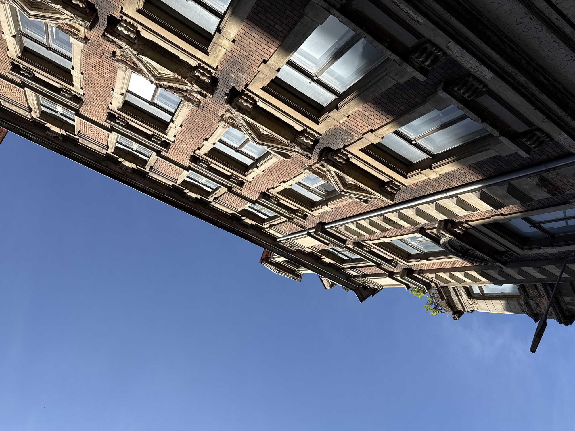 The image shows a building facade with brick and stone detailing, viewed from a low angle against a clear blue sky. The building has multiple windows, many of which reflect the sky. The architectural style suggests an older European building, possibly residential or commercial. The lighting indicates it is daytime, with sunlight casting shadows on the facade. There are no people or vehicles visible in the image. The scene appears to be an urban environment. Based on the architectural style, the building is likely located in Görlitz, Germany, a city known for its well-preserved historic architecture.