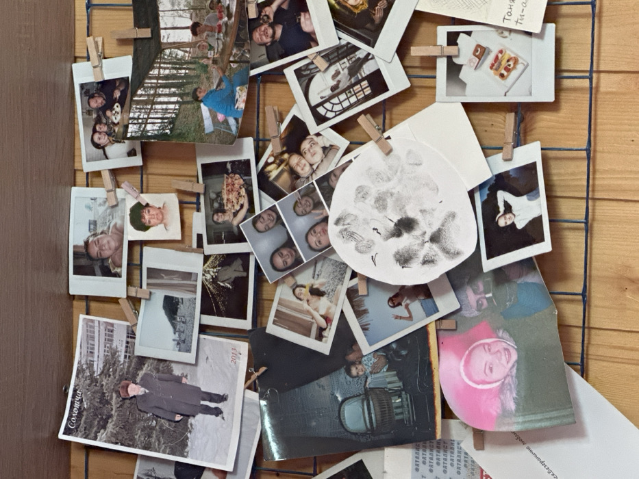 A vertical photo display board, constructed from a light brown wooden surface with an overlaying dark blue grid, presents a dense arrangement of personal photographs. Approximately 30-40 images of various sizes, predominantly small, instant-print style with white borders, are affixed to the grid using numerous small wooden clothespins. The scene is located in 3-Y Uchastok, Russia.
Notable photographs include a large vertical print in the upper-left depicting an older man in a dark coat and hat standing in a snowy landscape with a building; this photo bears the Russian text "Солотча" and the year "2013." Adjacent images feature a close-up portrait of a young woman and a beach scene. Further right, a group of people is shown gathered around a table with food, and another larger print displays individuals seated at a wooden table in a wooded outdoor setting under a tarp.
Central to the display is a prominent circular white paper bearing multiple dark gray fingerprints or smudges. Surrounding this are various photos portraying individuals and groups, including young women taking selfies, casual portraits, and scenes depicting social gatherings. Specific images include a vintage-style photo of a baby in a wicker pram, a smiling young woman in a pink hat, a woman posing on a beach, an architectural interior, and a close-up of miniature desserts. Partially visible Russian text, including "ФАТАЛИСТ" (Fatalist) and a handwritten note starting with "Таня" (Tanya), appear on papers attached to the lower portion of the board. The collection collectively documents personal memories and events.