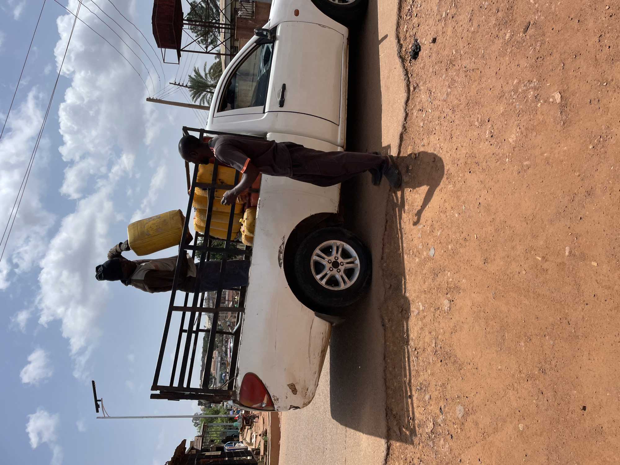 A white pickup truck is parked on the side of a dirt road, presumably in Jos, Nigeria. The truck bed is filled with yellow jerrycans, likely carrying water. Two men are interacting with the jerrycans, one pouring liquid from one can into another. The surrounding environment includes buildings, power lines, and other signs of a densely populated urban setting.