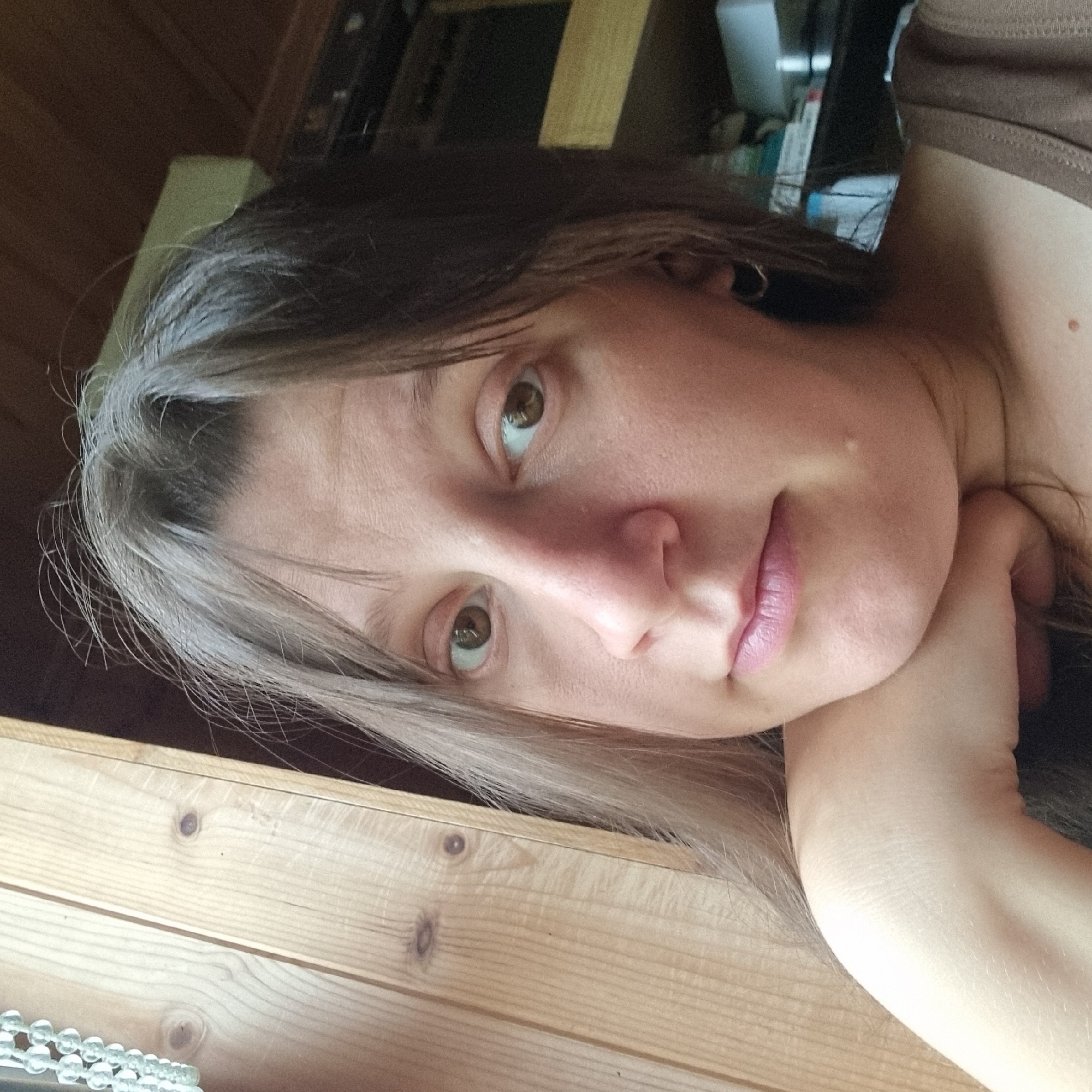 A close-up, chest-up, selfie-style photograph shows a woman with fair skin and light brown hair. She is leaning her head on her left hand, with her chin resting on her hand. Her eyes are open and looking towards the camera. Her mouth is closed in a neutral expression. The background consists of wooden paneling on the left and a dark, indistinct area on the right, possibly shelves or furniture. The lighting is natural and comes from the left side of the frame, casting a slight shadow on the right side of her face. There is a beaded string hanging on the wooden paneling in the upper left corner of the image. The scene does not provide any specific details about the city of Dunnae-myeon, South Korea.