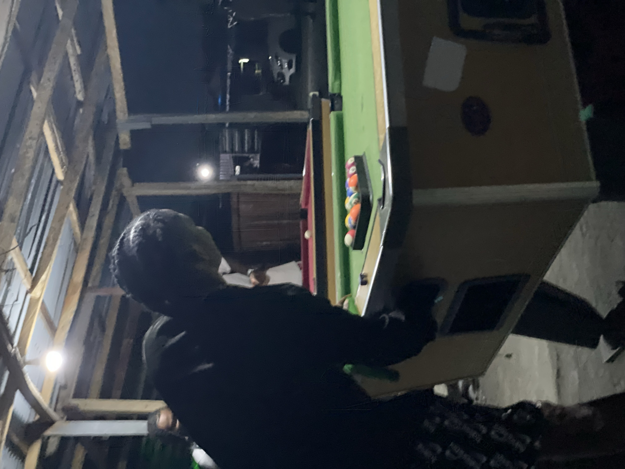 The image depicts an indoor scene at night or in low light, focusing on a person playing billiards. In the foreground, a person wearing a dark long-sleeved top is seen from behind, positioned as if to take a shot at a pool table. Their head is turned towards the table. Another person's head is partially visible further back, near the table.
The main object is a pool table with a green felt surface, oriented vertically in the frame from the camera's perspective. A rack of colorful billiard balls, including solids and stripes in orange, blue, red, and purple, is visible at one end of the table. The table has a light-colored (beige or light brown) frame, with a white square sticker and a dark circular mark on its side.
The setting appears to be an informal interior space, possibly a game room or a common area in Ado, Nigeria. The ceiling features prominent wooden beams or trusses, suggesting a rustic or unfinished structural style. The scene is illuminated by two bright, artificial light sources – one in the upper left and another in the central background – casting light against the dark surroundings.
The activity taking place is a game of pool. The overall mood is casual and focused on the game. No specific emotions are discernible from the visible subjects. There is no visible text in the image.