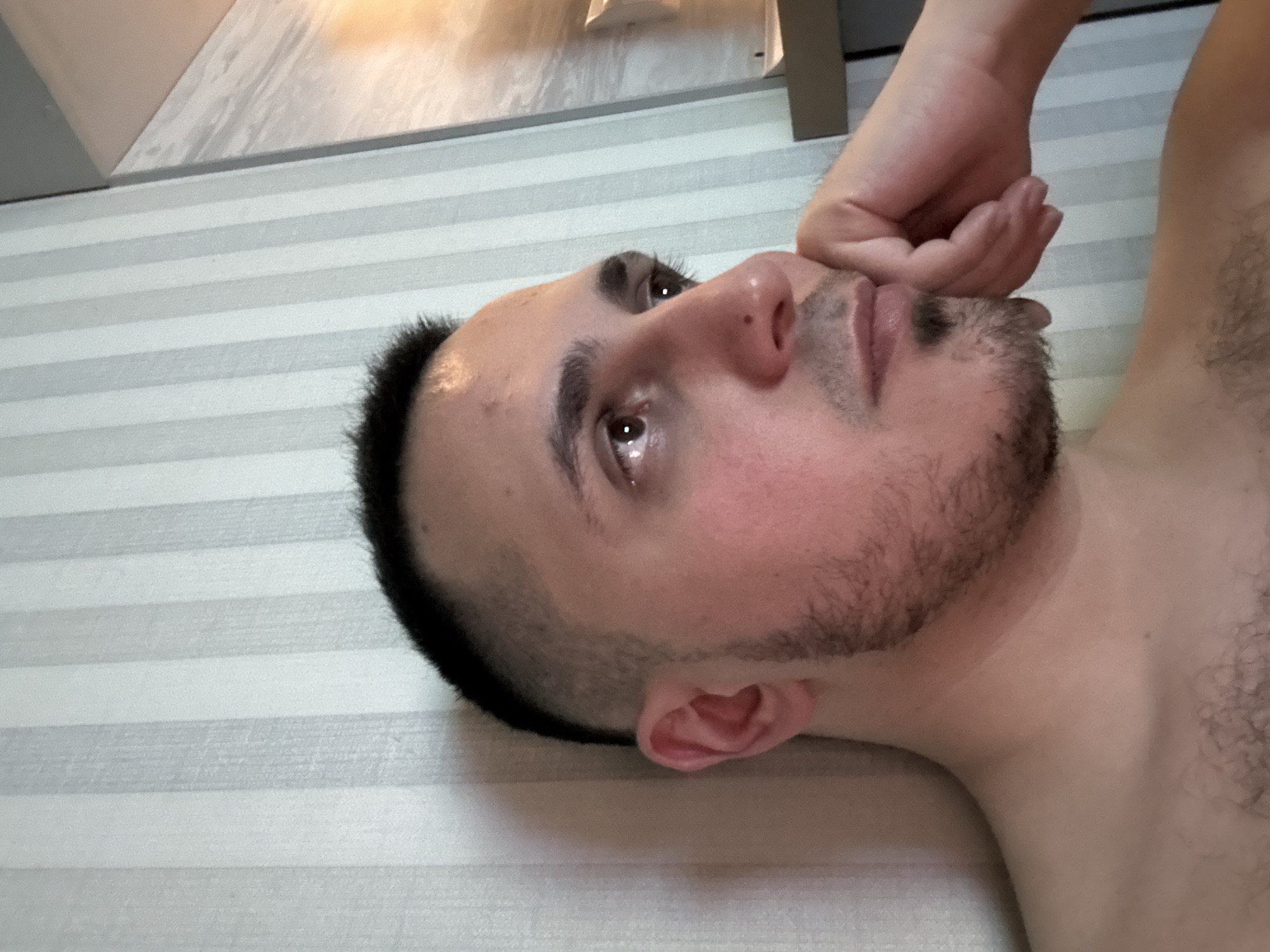 A close-up shot features a man lying down indoors, viewed from a slightly high angle, with his head turned to the right. His eyes are open and directed upwards, appearing slightly wet or reflective. He has short, dark hair with faded sides and a short beard or stubble on his face. A small dark mole is visible on the bridge of his nose. His left hand is positioned near his chin and mouth, with fingers slightly curled, suggesting contemplation or pensive thought.
The man's skin is fair, and there is visible body hair on his chest and shoulder area, particularly on the right side of the frame. The background consists of a surface with horizontal stripes, alternating between light and dark grey or white, resembling bedding or a wall covering. Above the striped surface, a blurred, light-colored ceiling or wall is visible, with hints of a rectangular object or furniture in the upper left and right corners. The overall lighting suggests an indoor setting, and the man's expression and posture evoke a sense of introspection or weariness. No text is visible in the image.