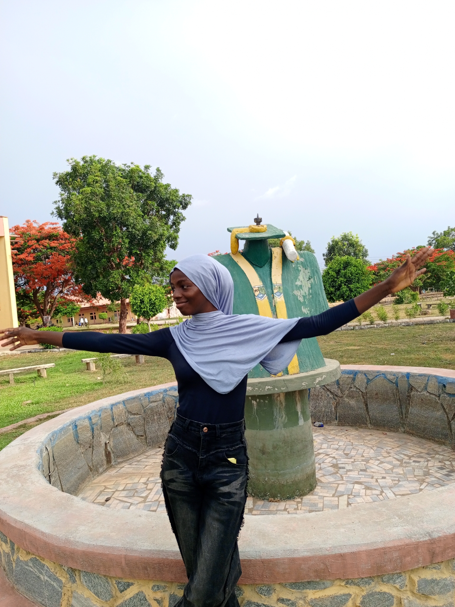 A medium, outdoor shot depicts a young woman standing in front of a statue within a round, raised, tiled platform with her arms outstretched. The woman has dark skin, a slim build, and is wearing a dark long-sleeve shirt, dark jeans, and a light blue hijab. She is smiling and looking to the left of the frame.
The statue behind her is of a graduation gown and mortarboard, painted in green, yellow, and white. The statue stands on a cylindrical base. The tiled platform surrounding the statue is built with stone on the outside.
In the background, there is a grassy area with trees and bushes. Some of the trees have red flowers. There is a building visible in the left background. The sky is overcast.
The location is in the city of Unknown, Unknown.
