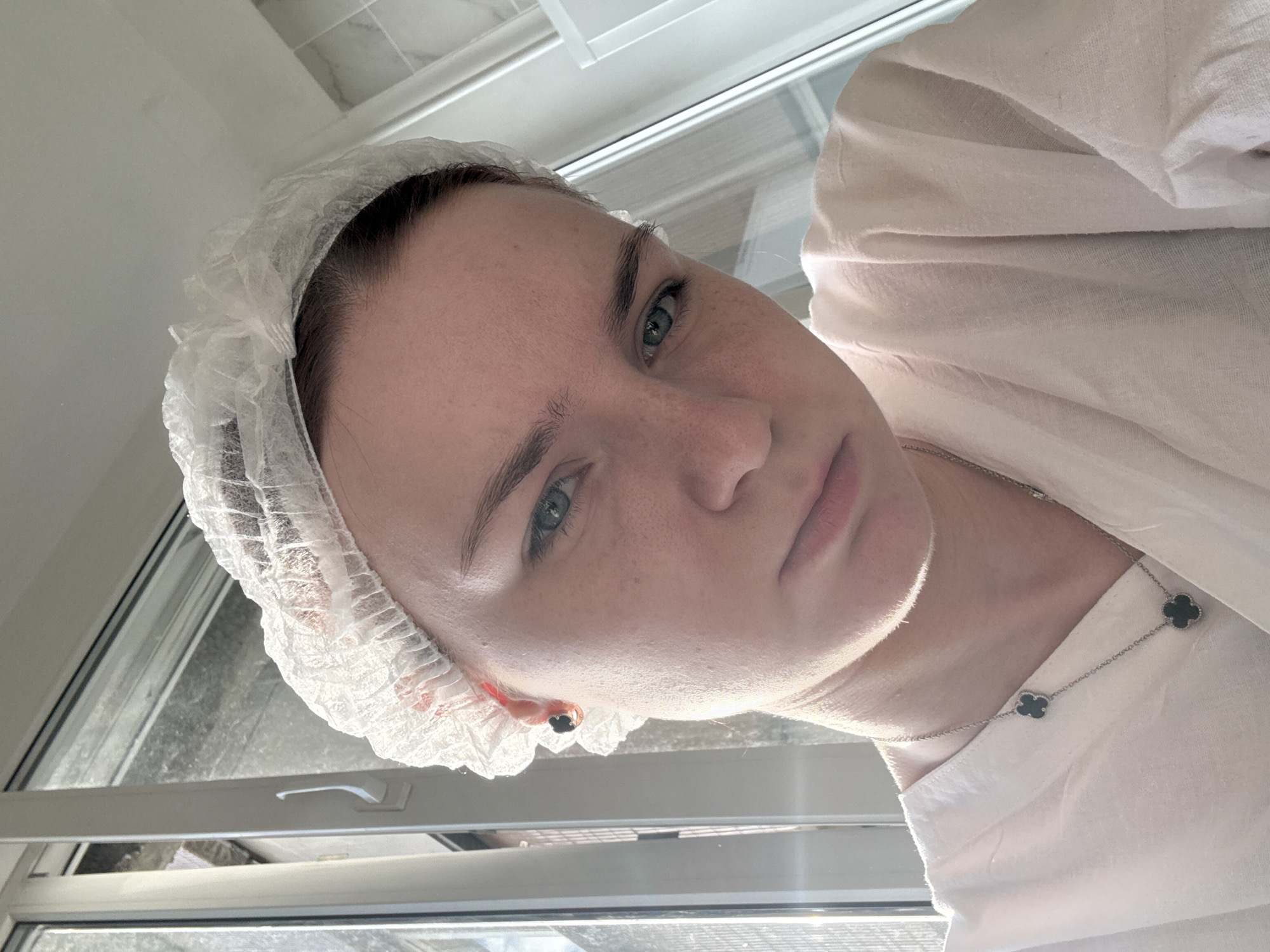 A close-up, selfie-style shot features a young woman wearing a white disposable hair net and a pale pink smock. She has light blue eyes, fair skin with freckles, and dark eyebrows. A dark stud earring is visible in her left ear. She is also wearing a silver necklace with two four-leaf clover charms. The background suggests an indoor setting with a window showing a blurred outdoor view. The lighting is bright, casting shadows and highlighting the woman's facial features and attire.
