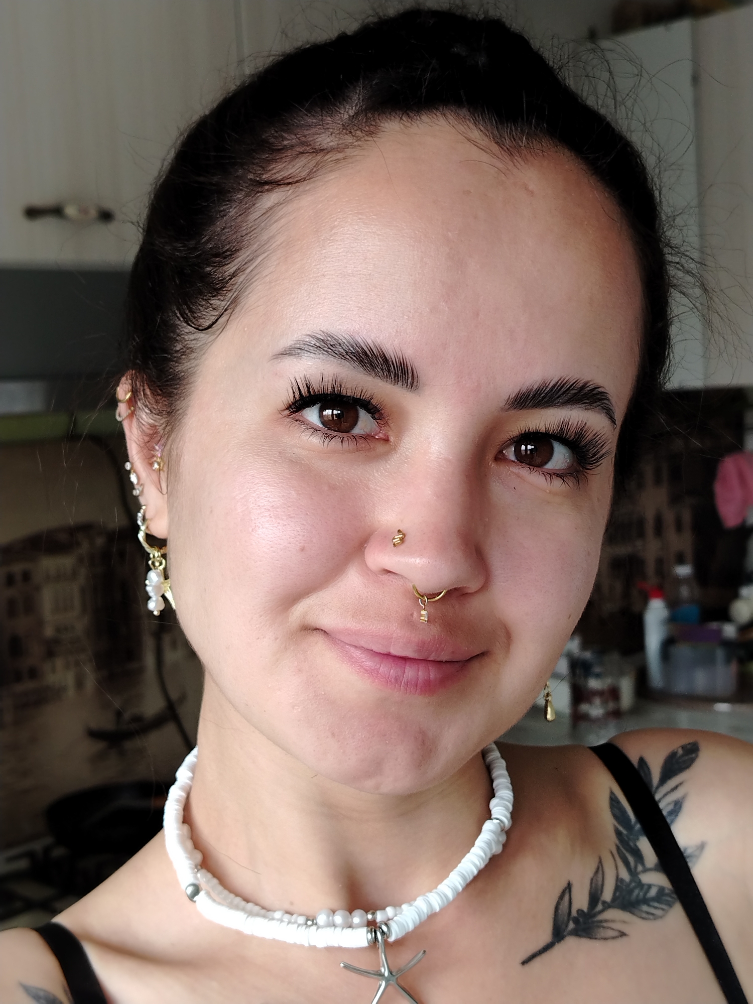 A medium shot portrait shows a young woman with dark hair pulled back, looking at the camera with a slight smile. She has noticeable makeup, including dramatic false eyelashes and well-defined eyebrows. Her left ear is adorned with multiple earrings, including a pearl and star charm. She has a nose ring and a septum ring. A white beaded necklace with a starfish pendant rests on her collarbone. A tattoo of a leafy branch is visible on her right shoulder. The background is blurred, suggesting an indoor setting with kitchen cabinets and a wall. The lighting indicates a daytime scene, possibly with natural light. There are no visible landmarks or indications of the specific location in Noviy, Russia.