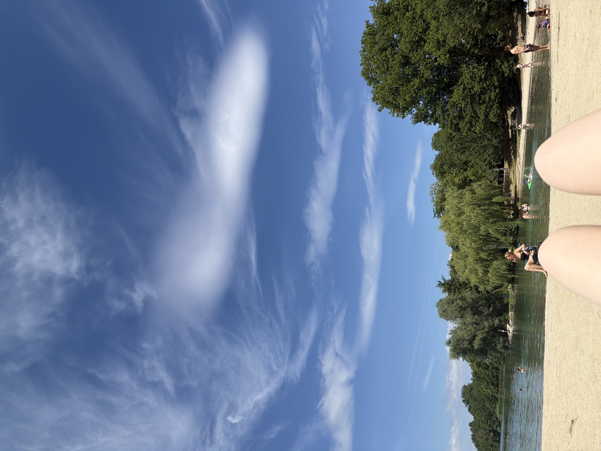 The image depicts a sunny day at a beach on the Robertsau Island in Strasbourg, France. The foreground is dominated by the legs of a person lying on the sand, facing away from the camera. The beach gives way to a body of water where several people are swimming and playing. In the background, a lush green forest surrounds the water, with tall trees creating a natural barrier. The sky above is a brilliant blue, dotted with wispy white clouds, indicative of fair weather. Several individuals are visible in the water, some appearing to be swimming, while others are closer to the shore. A woman is seated on the sand in the mid-ground, facing the water. The overall scene conveys a sense of leisure and relaxation on a warm summer day.