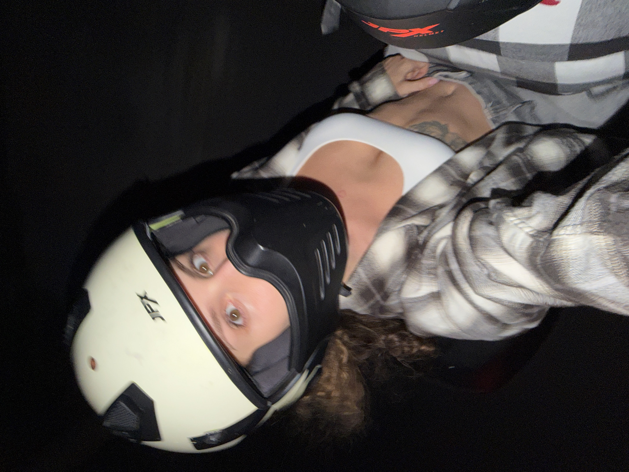 The image is a close-up, vertically oriented shot featuring a person wearing a motorcycle helmet and casual attire. The subject, likely female, has light brown, wide-open eyes looking directly at the camera. They are wearing a cream-colored motorcycle helmet with black accents and the "JFX" brand logo visible on the side. Underneath the helmet, a black mask-like piece covers the lower part of their face.
The person is dressed in a black and white plaid flannel shirt, worn open over a plain white top, possibly a tank top or sports bra. A small tattoo is visible on their upper chest. A portion of dark, curly hair can be seen emerging from beneath the helmet. In the upper right corner, part of another black helmet with red "FX HELMET" branding is visible.
The background is almost entirely dark, suggesting a nighttime setting or a dimly lit environment. The presence of two helmets implies a context related to motorcycling or scooter riding. The overall scene captures a moment of someone either preparing for or posing with their riding gear. The location context is North Kuta, Indonesia.