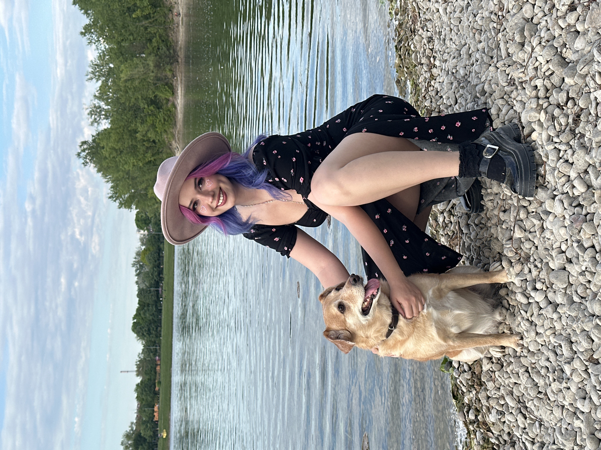 An outdoor, eye-level shot captures a young woman and a dog sharing a tender moment by a body of water in Zagreb, Croatia. The woman, with vibrant purple and pink hair and a light brown wide-brimmed hat, kneels on a pebbled shoreline. She wears a black dress adorned with small pink floral patterns and chunky black platform boots. Her face is turned towards the camera, beaming with a wide smile, and she has subtle heart-shaped glitter on her cheekbones, adding to her cheerful demeanor. Her right hand gently rests on the back of a light brown dog, which is sitting contentedly beside her. The dog looks up at her with its tongue playfully hanging out, a dark collar visible around its neck, reflecting its happy engagement.
To the left of the pair, the calm water stretches, showing gentle ripples on its surface. Beyond the water, a verdant treeline marks the distant bank, hinting at a natural setting. Above, a soft, cloudy sky suggests it is daytime, possibly late afternoon, with diffused light illuminating the scene. The overall impression is one of warmth, affection, and leisure, as the woman and her canine companion enjoy their time together by the water. There is no visible text in the image.