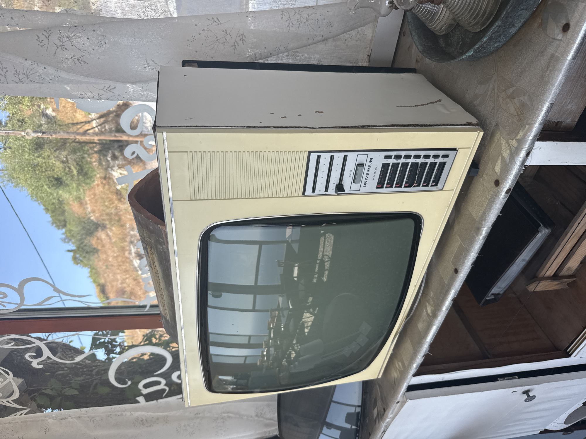 The image shows an old, beige Universum television set placed on a shelf in front of a window. The television has a dark screen that reflects the interior of a room, possibly a restaurant or cafe, with tables and chairs visible. Through the window, a sunlit hillside with trees and a clear blue sky can be seen. White curtains with a floral pattern hang on either side of the window. The shelf is covered with a textured, patterned material and appears to be part of a larger piece of furniture with an open compartment below. The setting appears to be in Λάκωνες, Greece, as indicated by the name on the building behind the window. There are no people or animals visible in the image.