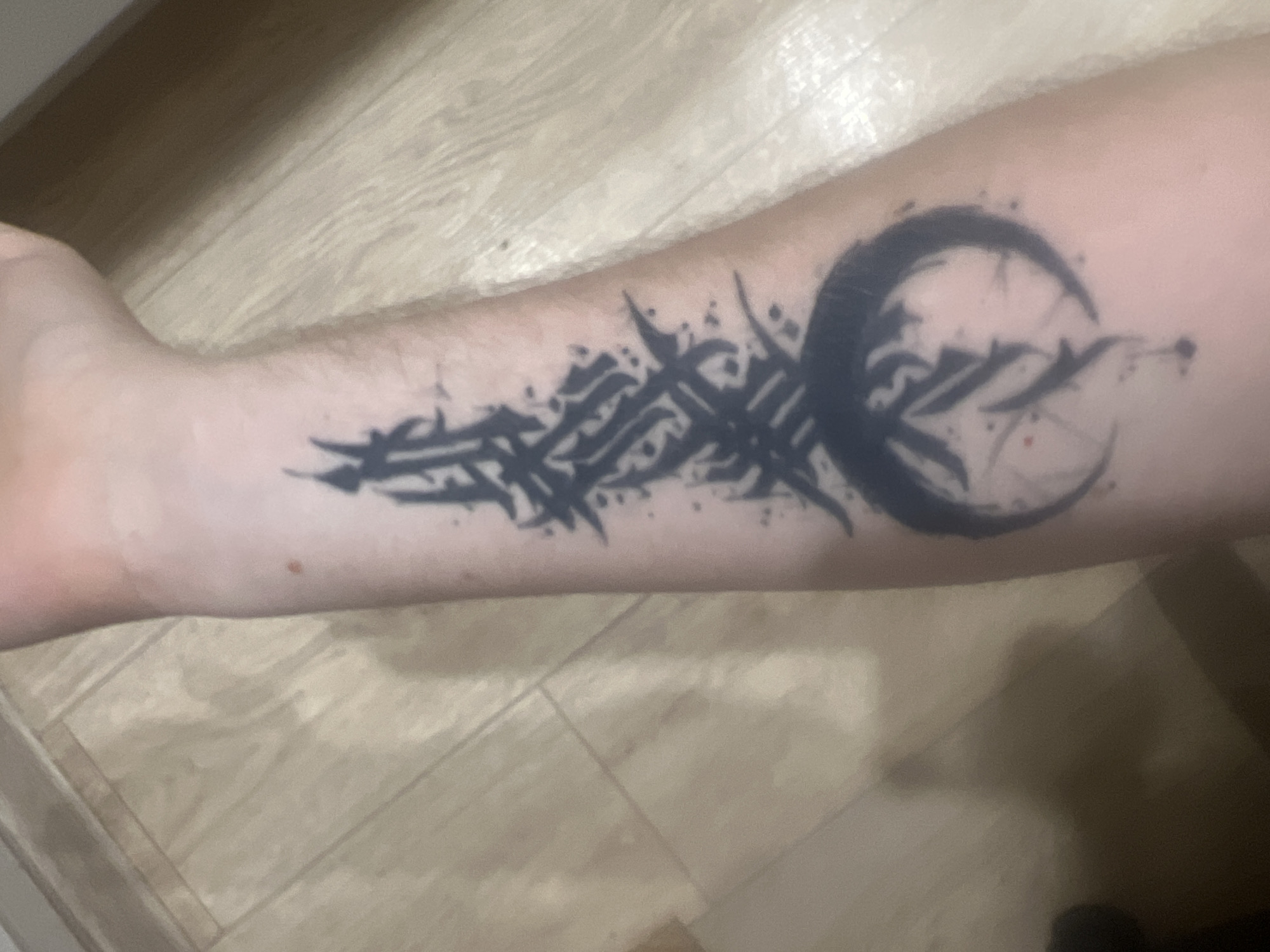 The image shows a close-up of a person's forearm with a black tattoo. The tattoo is abstract and features sharp, angular lines and splatters, resembling Gothic or tribal art. It appears to be a design with a crescent moon shape integrated into it, along with other intricate patterns. The tattoo covers a significant portion of the forearm. The skin tone is fair, and there are a few minor blemishes or moles visible. The forearm is resting on a light-colored, wooden surface, possibly a table or floor, with visible grain patterns and lines suggesting planks. The lighting appears to be artificial, casting a soft, even illumination on the subject and background. The overall mood conveyed by the image is neutral, focusing on the details of the tattoo. No specific time of day, weather, or emotions are explicitly discernible. The location context is provided as Saratov, Russia, but no visual cues in the image directly confirm this. There is no visible text in the image.