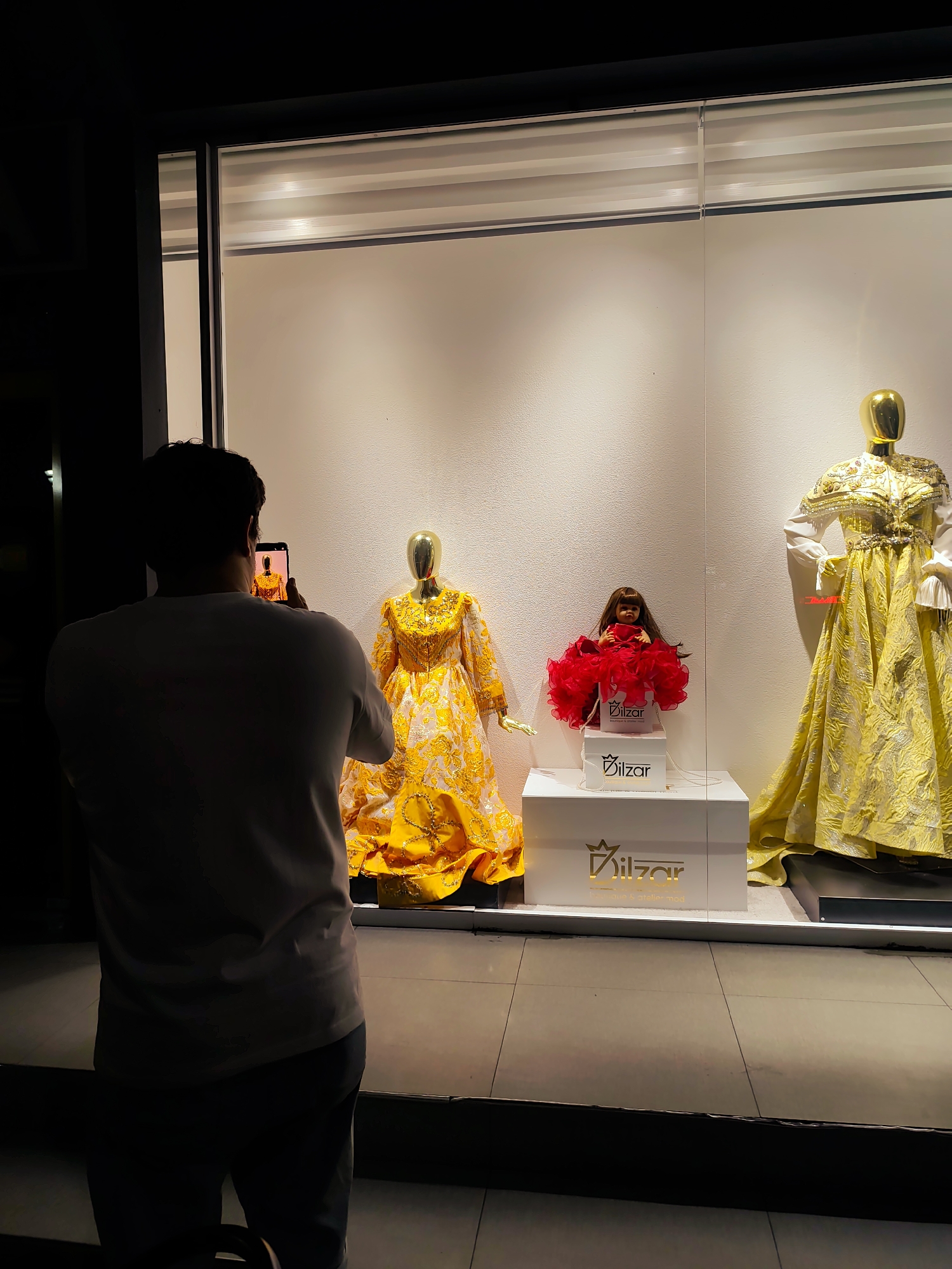 In Fergana City, Uzbekistan, a person stands in silhouette, facing away from the camera and holding a smartphone to take a picture of a store window display. The display features three mannequins dressed in ornate, yellow gowns. The left mannequin is partially visible, wearing a yellow gown with intricate patterns. The center features a doll in a bright red dress, placed on a white box with gold lettering that reads "Dilzar". To the right, a mannequin is adorned in a elaborate yellow gown with gold embellishments and white sleeves. The display is well-lit, with the interior of the store having a clean, modern aesthetic, suggested by the white wall and horizontal blinds. The floor in front of the display is tiled. The overall scene depicts a street-level view of a boutique or clothing store, captured at night.