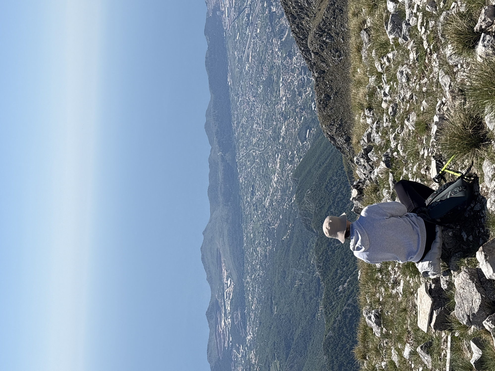 The image depicts a person sitting on a rocky, grassy cliff overlooking Mali Mikulići, Montenegro. The person wears a light gray hooded sweatshirt and a tan hat. A dark backpack rests behind them, with yellow trekking poles attached. The background shows a vast landscape, including mountains and a city below, under a clear blue sky. The perspective is from above, looking down at the person and the surrounding environment.