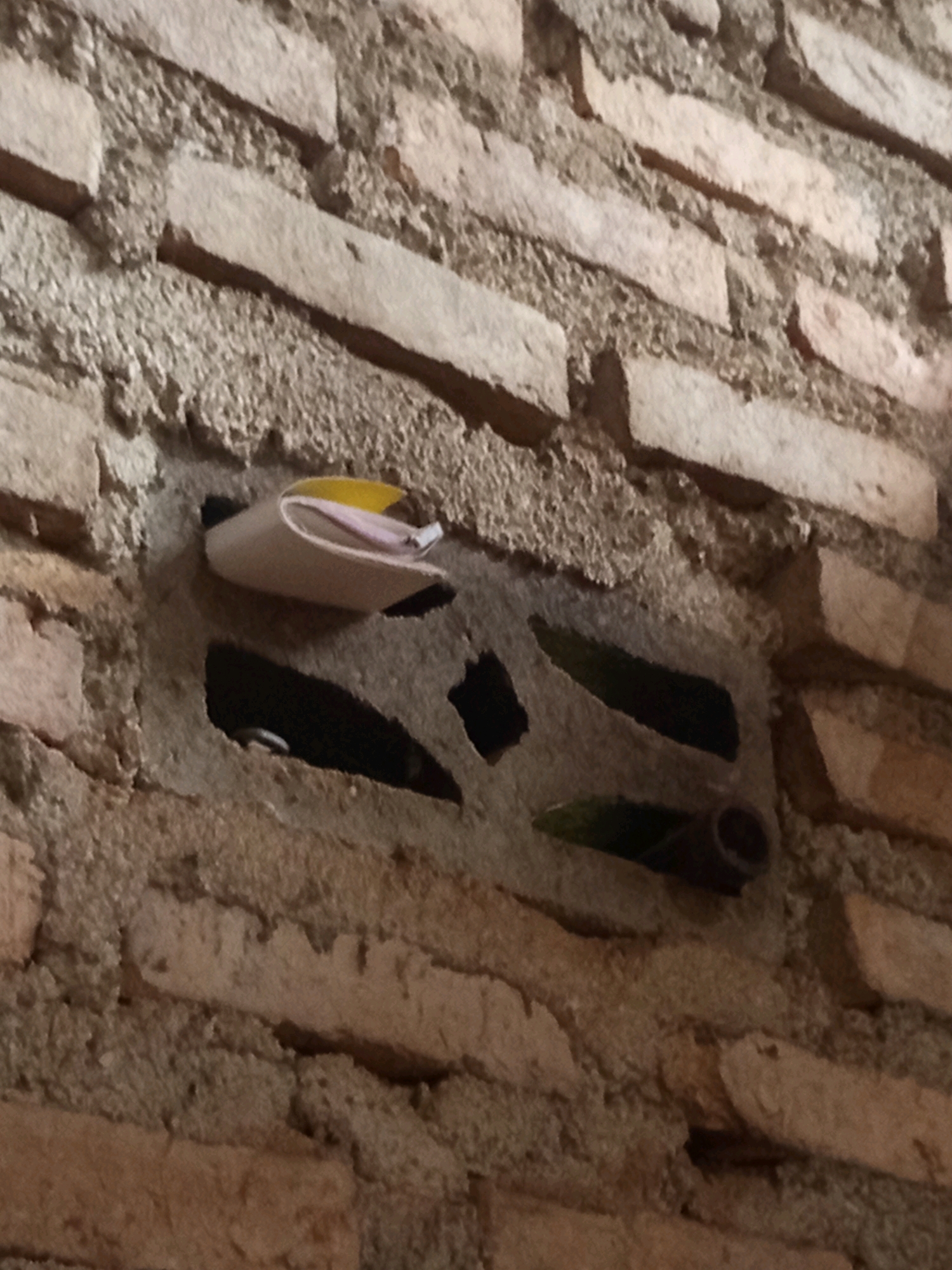 The image shows a close-up view of a brick wall with a small, light-colored object tucked into a hole. The object appears to be a folded piece of paper or a small container, with hints of yellow and pink visible. The brickwork is rough and aged, with visible mortar lines. There are other openings in the wall, some dark and some with glimpses of what might be pipes or other objects. The scene is located in the city of Karang Sari, Indonesia.