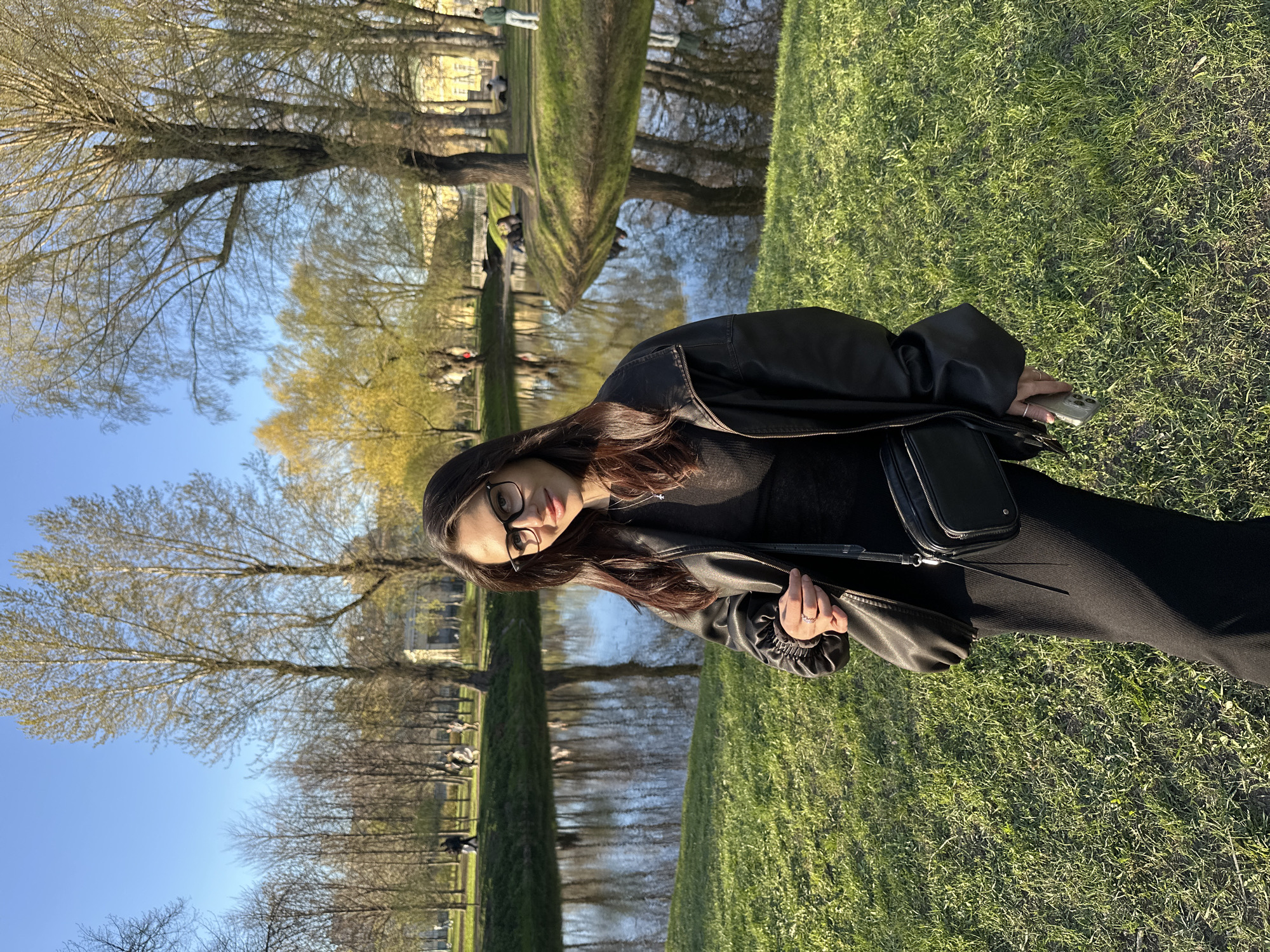 The image shows a woman standing on a grassy area. She is wearing glasses, a black top, a black leather jacket, and black pants. She has long brown hair. The woman is holding a cell phone in her right hand and the strap of a black purse with her left hand.
In the background, there is a body of water, possibly a canal or river, lined with trees. Beyond the water, there is a building. People can be seen in the background, some sitting on the grass.
The scene is in Saint Petersburg, Russia.