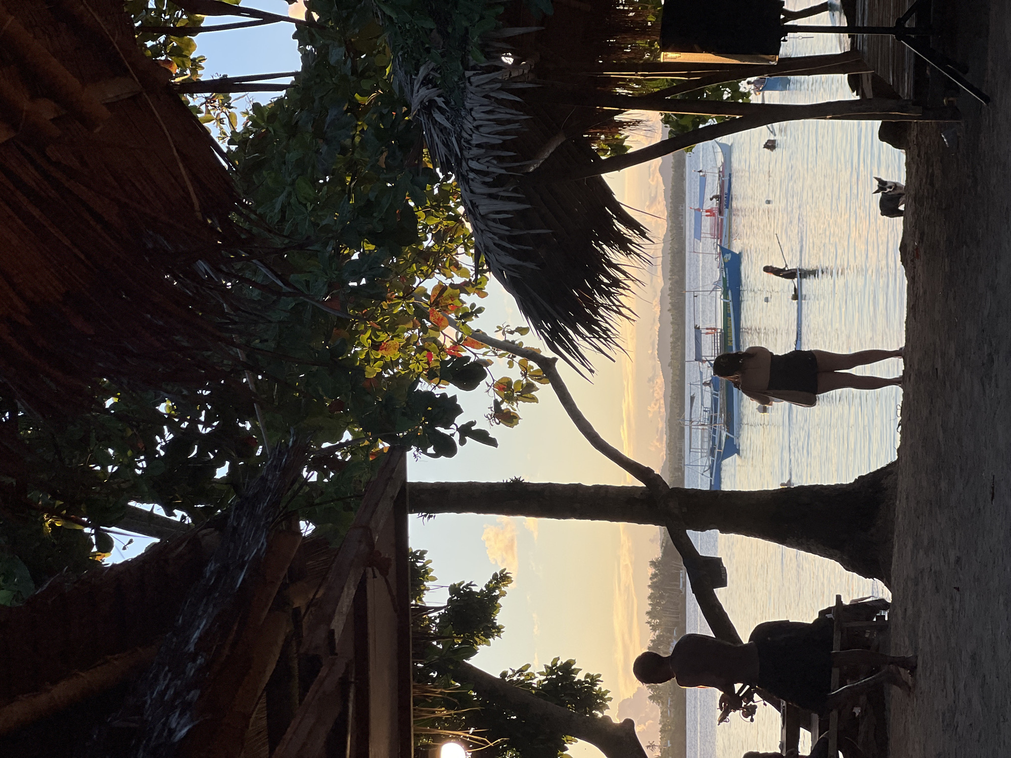 A silhouette of a man stands on a sandy beach in front of a body of water, holding a phone in his left hand. He is wearing a dark skirt and is barefoot. To his right, a woman with dark hair stands facing the water, wearing a black top and shorts, and carrying a beige bag over her shoulder. In the background, several boats are anchored in the calm, reflective water. The scene is framed by the thatched roofs and palm trees of a beachfront establishment, suggesting a resort or restaurant. The sky above is a soft gradient of yellow and blue, with scattered clouds, indicating either sunrise or sunset. In the foreground, a dog stands looking towards the camera. The overall impression is one of a serene tropical evening in General Luna, Philippines.