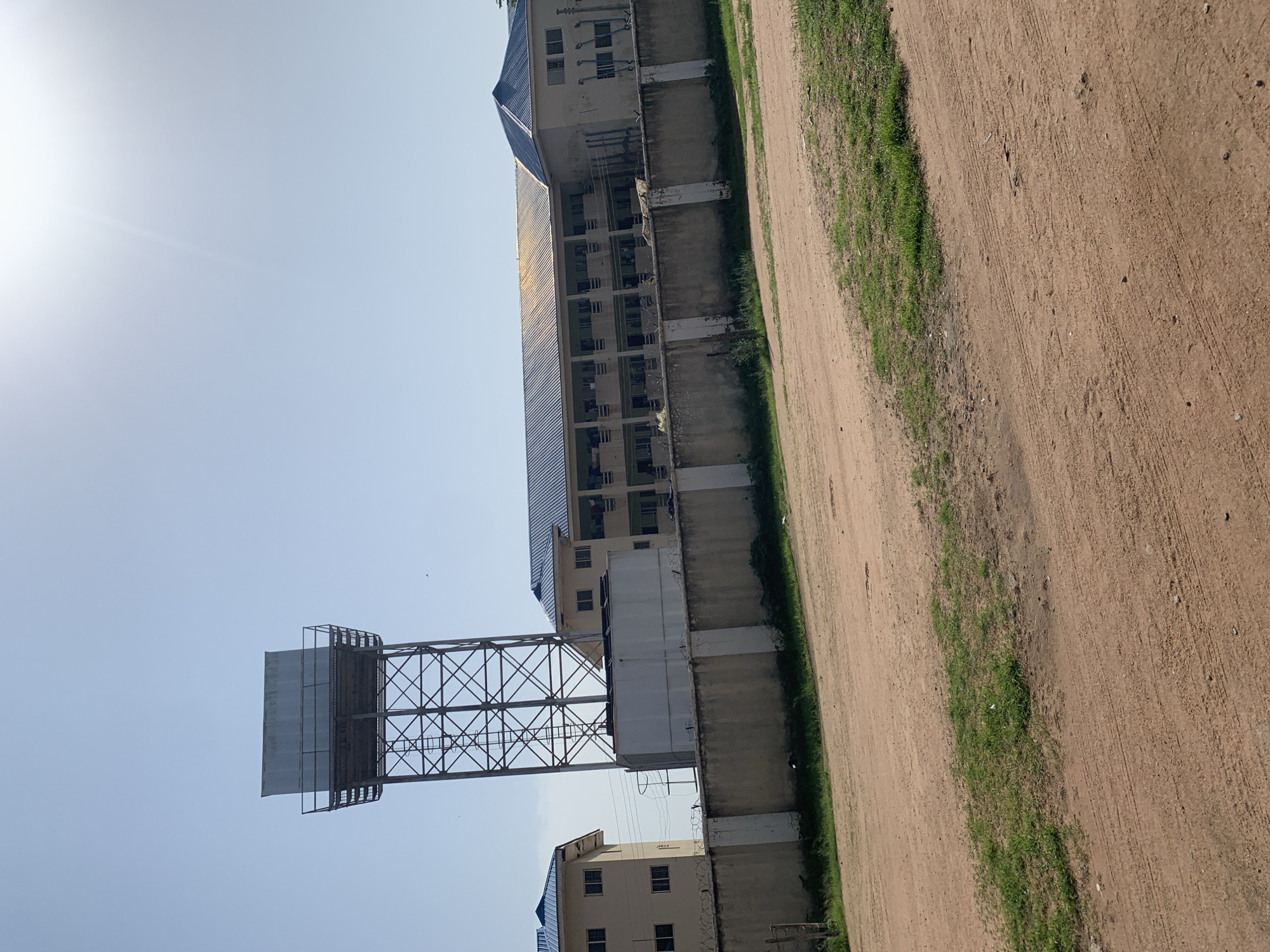 A wide shot captures a three-story building with a blue roof and numerous windows, characteristic of student housing or a dormitory, situated behind a concrete wall topped with barbed wire. Adjacent to this building, a taller structure with a similar blue roof is partially visible on the left. Dominating the left side of the frame is a tall, metal water tower supporting a large rectangular tank. The foreground features a dirt road with sparse grass patches. The sky is clear and blue, with the sun creating a glare in the upper right quadrant of the image. The overall setting suggests an academic or institutional campus in Jalingo, Nigeria.