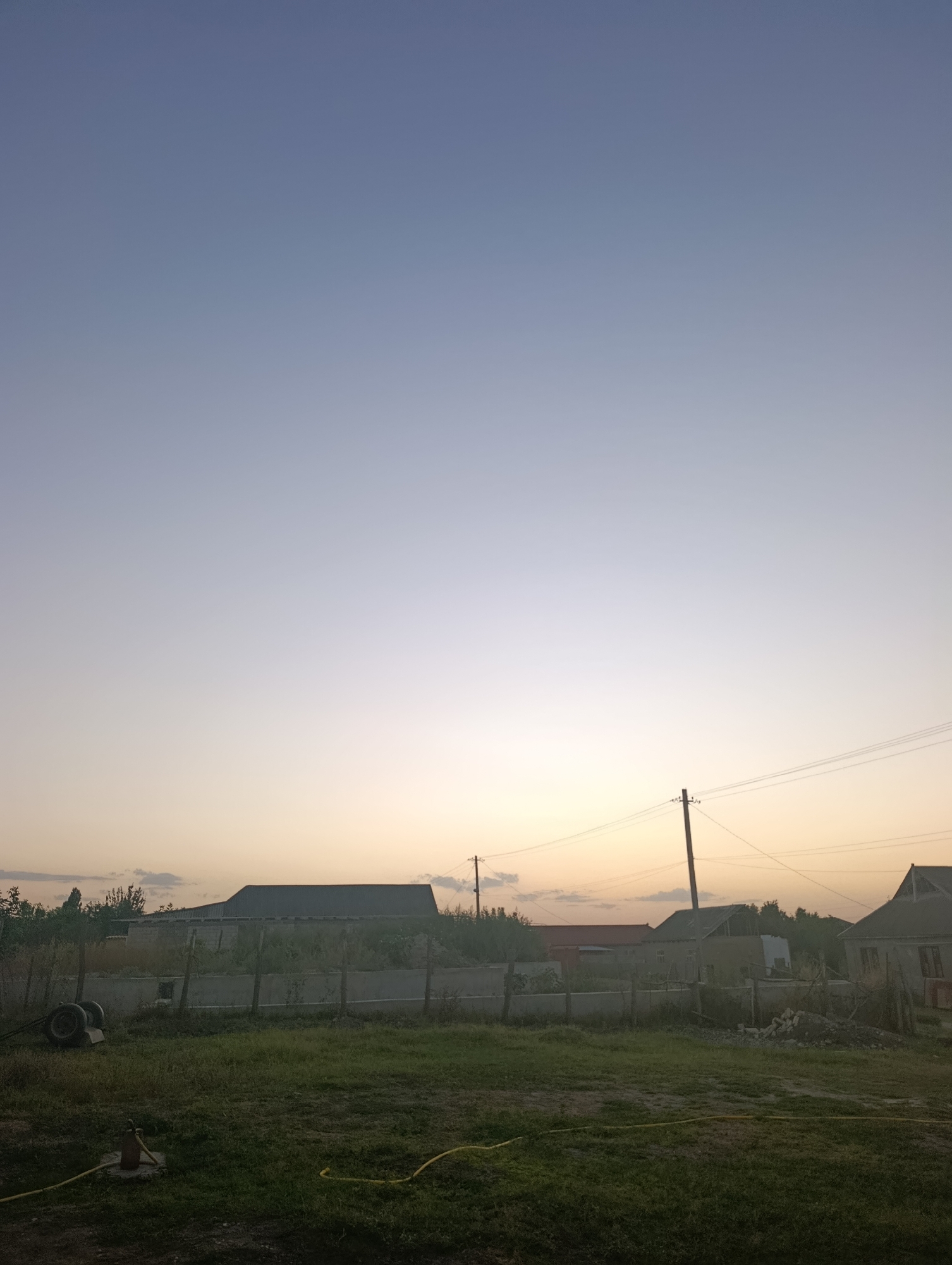 This image depicts an outdoor scene at dusk, likely in a rural or suburban area, with a vast, gradient sky transitioning from pale blue at the top to a soft yellow and orange hue near the horizon. In the midground, there are several structures including a long, dark-roofed building resembling a barn or workshop, and other smaller buildings with visible roofs and walls. A fence runs across the scene, separating the foreground from the buildings. A utility pole with wires is visible to the right of the frame. The foreground consists of a grassy area with some miscellaneous objects. On the left, there are two tires stacked on top of each other. A yellow hose is visible snaking across the grass in the lower portion of the image. The overall impression is of a quiet, domestic setting at the end of the day.