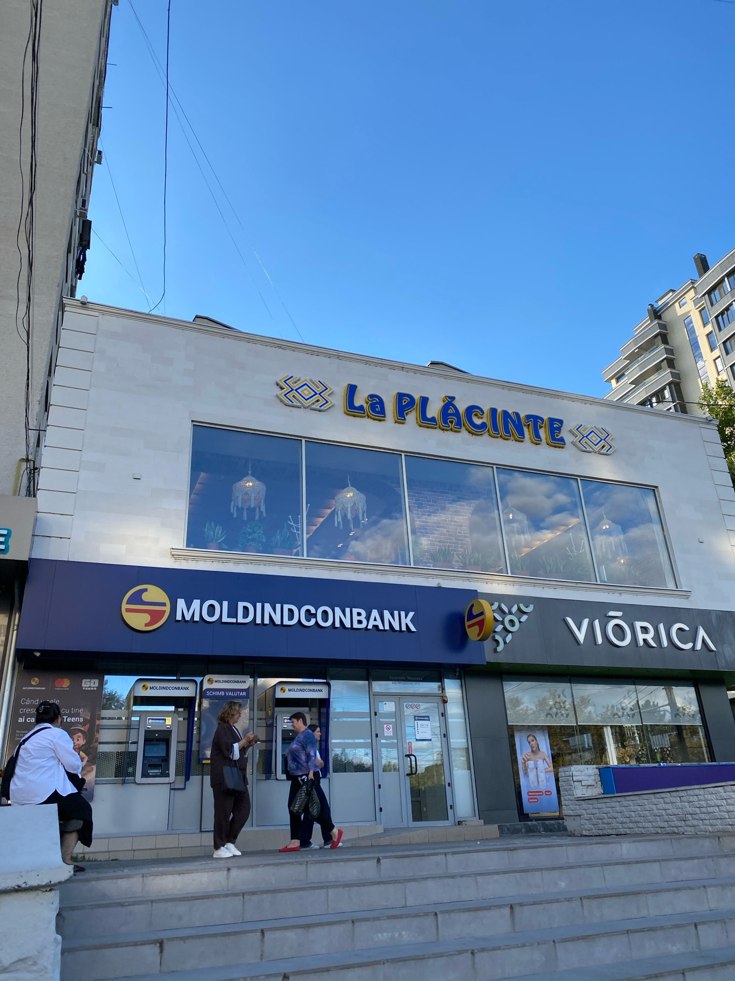 The image shows a street-level view of a building in Chișinău, Moldova, with storefronts and an ATM area. The building has a modern facade with a light-colored stone finish on the upper level, featuring the signage "La Plăcinte" in blue and yellow lettering, indicating a restaurant. Below this, a section of the building is dedicated to "MOLDİNDCONBANK," a bank, with ATMs visible. To the right of the bank, there is a storefront labeled "VIORICA," which appears to be a cosmetics store, as indicated by a poster featuring a woman and beauty products.
In the foreground, a set of concrete stairs leads up to the entrance of the building. Several people are present:
* On the left, a person in a white shirt and black pants is seated on the edge of a concrete structure, looking towards the ATM area.
* In the center, two individuals are standing near the ATMs. One person in dark clothing is looking at their phone, and another person in a patterned shirt and dark pants is also near the ATMs.
* To the right, two people are walking away from the ATMs. One is wearing a blue and purple top and dark pants, carrying shopping bags. The other person is wearing dark clothing.
The sky is clear and blue, suggesting daylight. The overall environment appears to be a commercial street in the city.