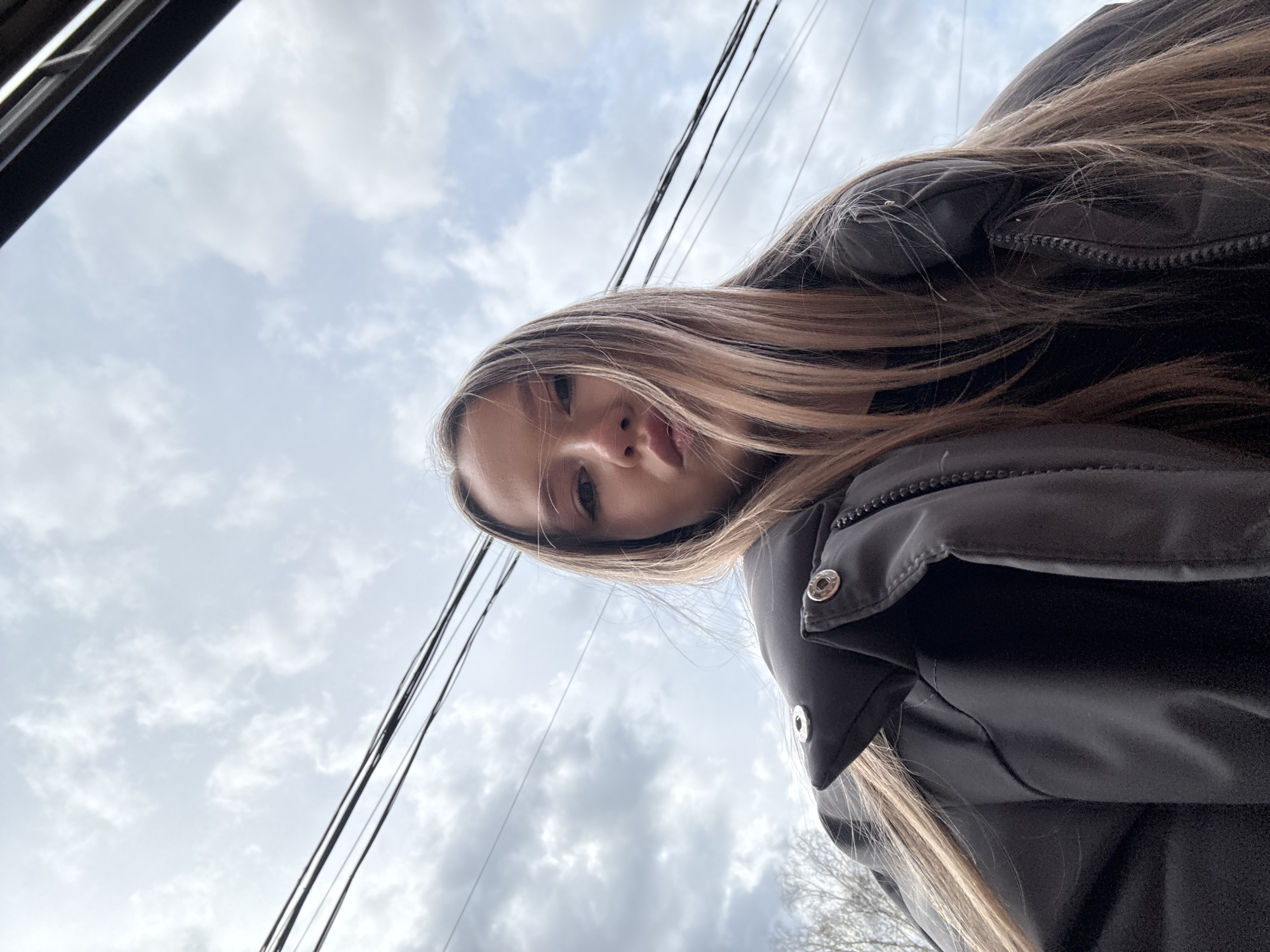 An upward-angled shot captures a young person, likely female, from a low perspective. Her face, showing her nose, mouth with slightly parted lips, and portions of her eyes and forehead, is visible in the center-left of the frame. Long, light brown hair cascades around her, partially covering her shoulders and flowing onto her jacket. She is dressed in a dark, possibly black, puffer-style jacket with visible metallic snap buttons and a zippered detail on the right side.
The background primarily features a cloudy sky, displaying a mix of lighter and darker grey clouds, indicative of an overcast day. Several dark utility lines, appearing as power or telephone lines, stretch across the sky in the upper right quadrant of the image. A dark, angled structure, possibly a window frame or part of a vehicle, is visible in the upper left corner. Faint bare tree branches can be discerned in the lower right portion of the background.
The image's perspective suggests a selfie or a photograph taken from below, looking up at the subject and the sky. The scene depicts an outdoor environment, likely in an urban or suburban setting within Фоки, Russia, during daytime under cloudy weather conditions. No visible text is present in the image.