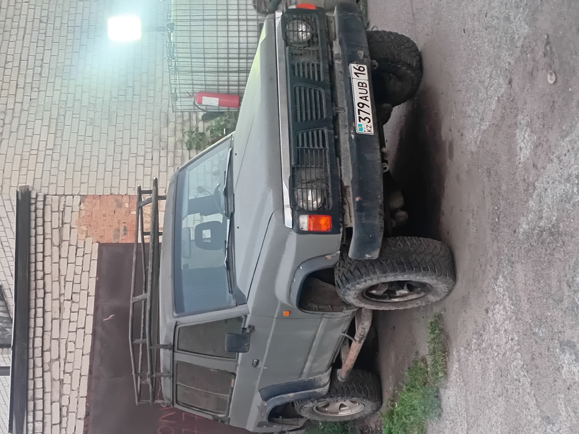 A weathered, grey, two-door SUV is parked on a cracked asphalt surface in front of a brick building in Semey, Kazakhstan. The vehicle has a roof rack, aftermarket fenders, and off-road tires. A license plate on the front bumper reads "KZ 379 AUB 16." A fire extinguisher is visible to the right of the vehicle, next to a large caged structure. A bright light illuminates the scene from the right, casting a haze. The building's brick facade is visible in the background, with a dark, corrugated metal door.