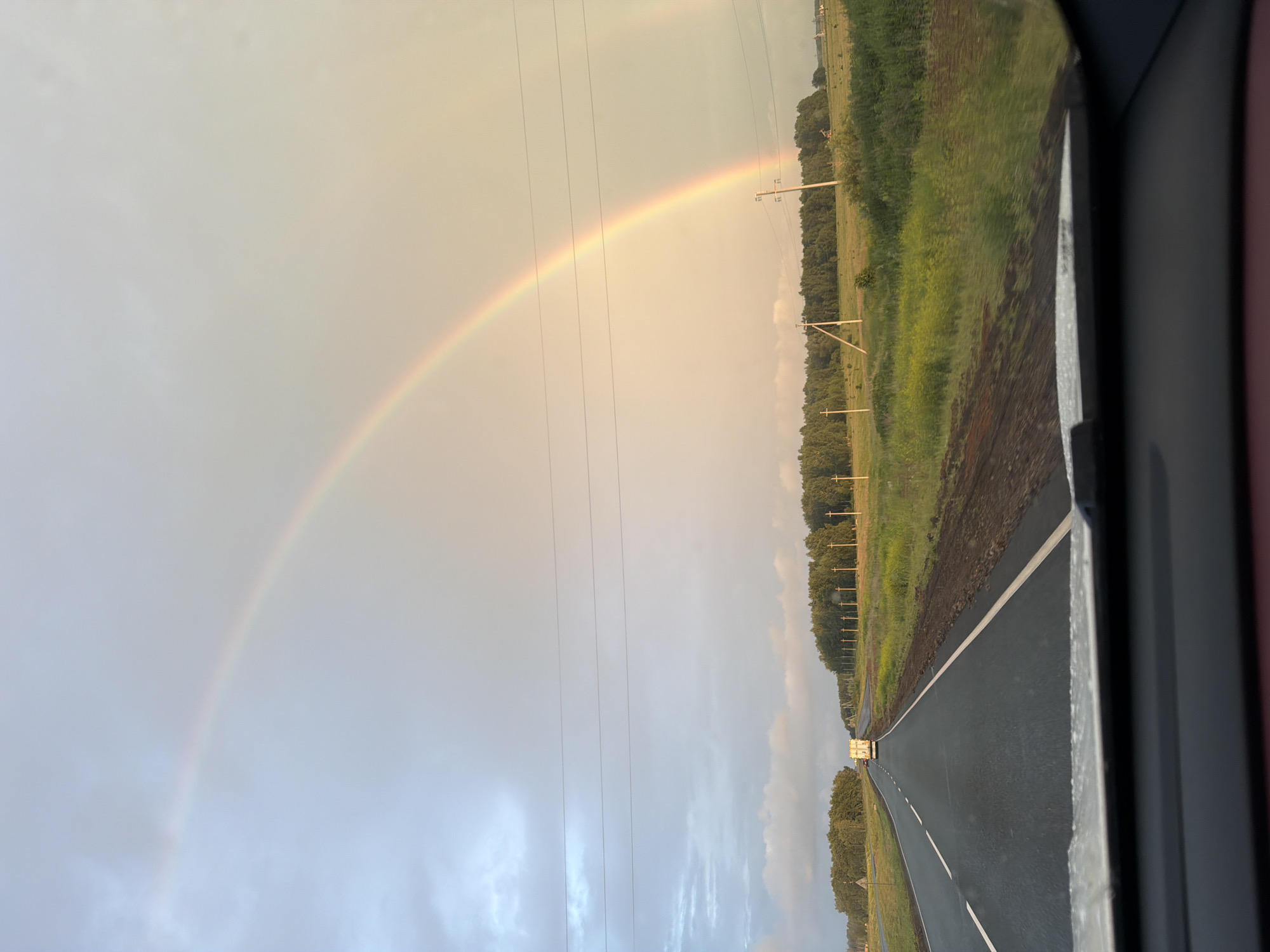 The image depicts a road with a double rainbow overhead, likely taken from inside a vehicle. The road stretches into the distance, bordered by green fields and a dense line of trees. Several utility poles are visible along the side of the road. A white truck is seen driving on the road ahead. The sky is overcast with the sun's light casting a warm glow, illuminating the rainbow. The scene is set in a rural environment, specifically in the city of Таймурзино, Russia, during daylight hours.