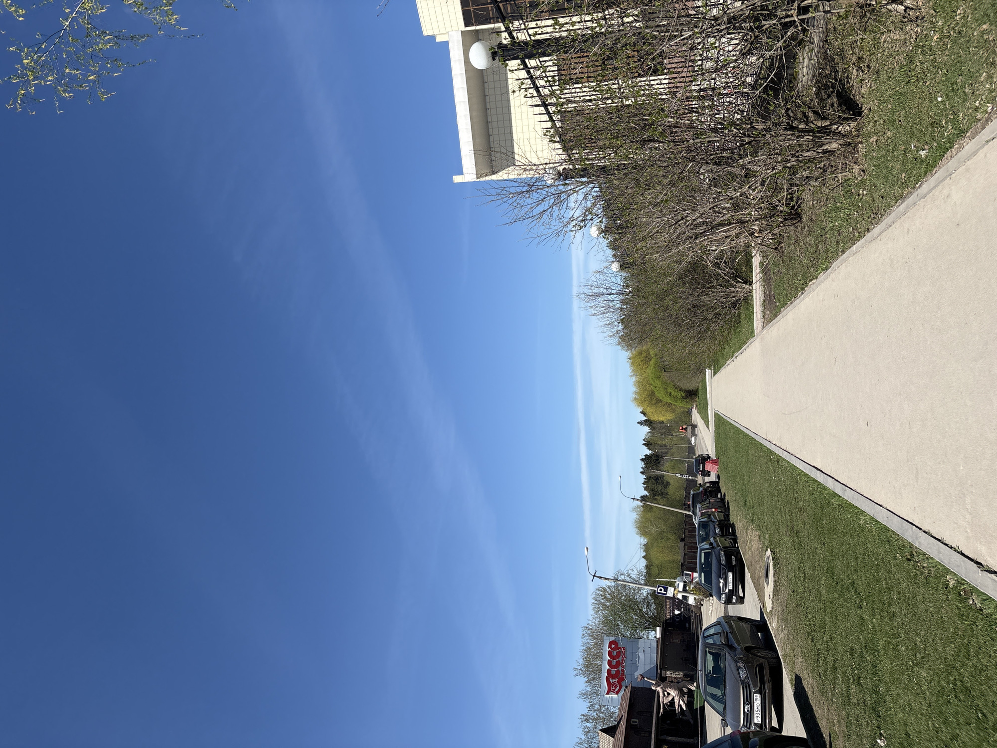 The image depicts a street scene in Perm, Russia, characterized by a clear blue sky and bright daylight. The composition is divided between the sky and a street view. A sidewalk runs along the right edge, bordered by a strip of green grass. A row of parked cars lines the street next to the grass. A building with a white facade and a fence is visible along the sidewalk in the upper right portion of the image. A sign with red letters is partially visible among the parked vehicles. Street lights line the street. Bare trees and emerging green foliage add to the urban landscape.