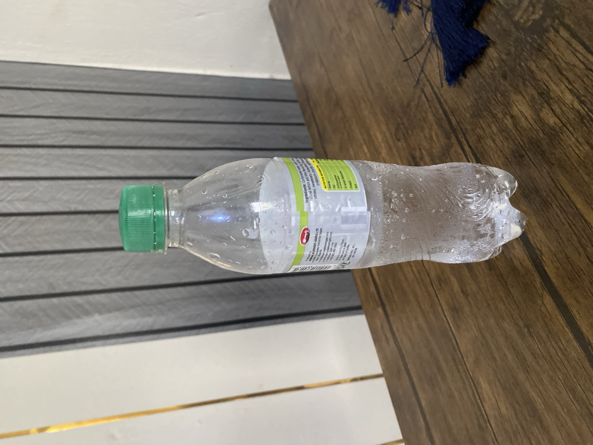 A clear plastic bottle filled with a carbonated beverage and condensation is centered on a wooden table. The bottle has a green cap and a label that is partially visible and appears to contain nutritional information and ingredients. The background consists of vertical gray blinds and a thin gold vertical element on the left. A navy blue fringe is visible on the right side of the table. The scene is indoors. The image does not provide any information about the location in the city of Jauro Yinu, Nigeria.