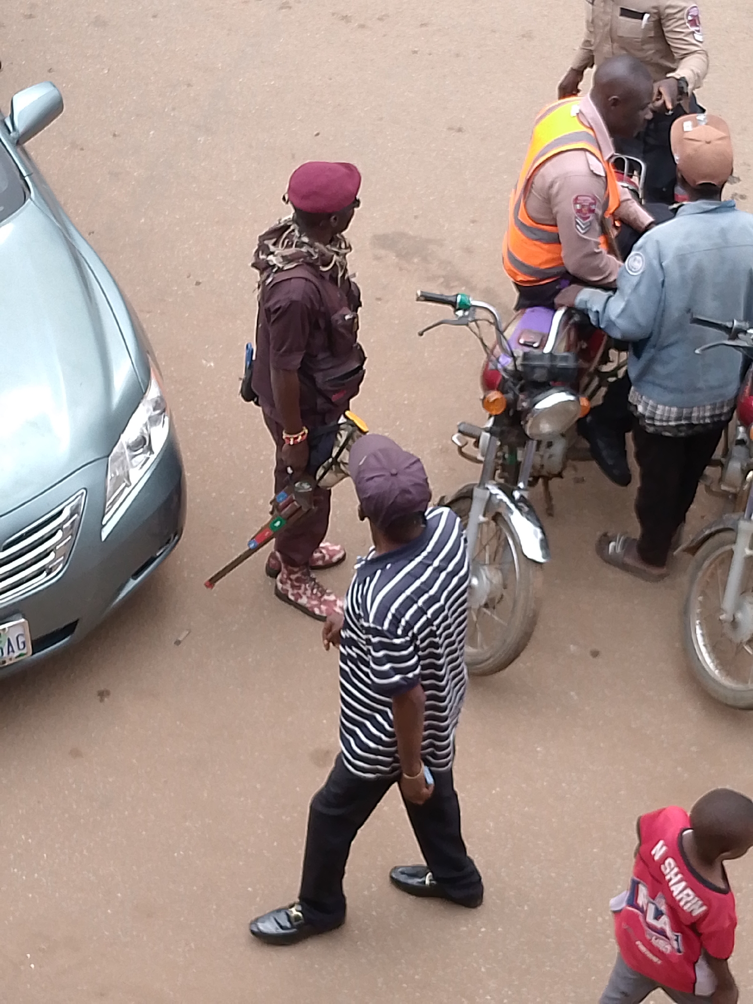 The image, taken in Bukuru, Nigeria, depicts an outdoor scene on a paved surface, likely a street or open area. Multiple individuals are present, some interacting with motorcycles.
In the foreground, a person wearing a striped long-sleeved shirt and black trousers walks away from the camera. They have a dark cap on their head and black shoes.
To the left, a silver car is parked. A person wearing a maroon beret and a maroon uniform stands near the car, holding what appears to be a rifle. They have a patterned scarf around their neck and patterned shoes.
Behind the car and to the right, three individuals are gathered around a motorcycle. One person, wearing an orange and tan reflective vest over a tan uniform, is seated on the motorcycle. Another person, wearing a tan uniform, stands behind the seated individual. A third person, wearing a blue denim jacket and a light-colored cap, is also near the motorcycle, facing the seated person.
Further to the right, another motorcycle is partially visible, and a young person in a red shirt with white lettering on the back is walking away from the camera. The red shirt has the words "N SHARIN" visible. The ground is a textured, light brown color, suggesting asphalt or a similar material. The lighting indicates it is daytime.