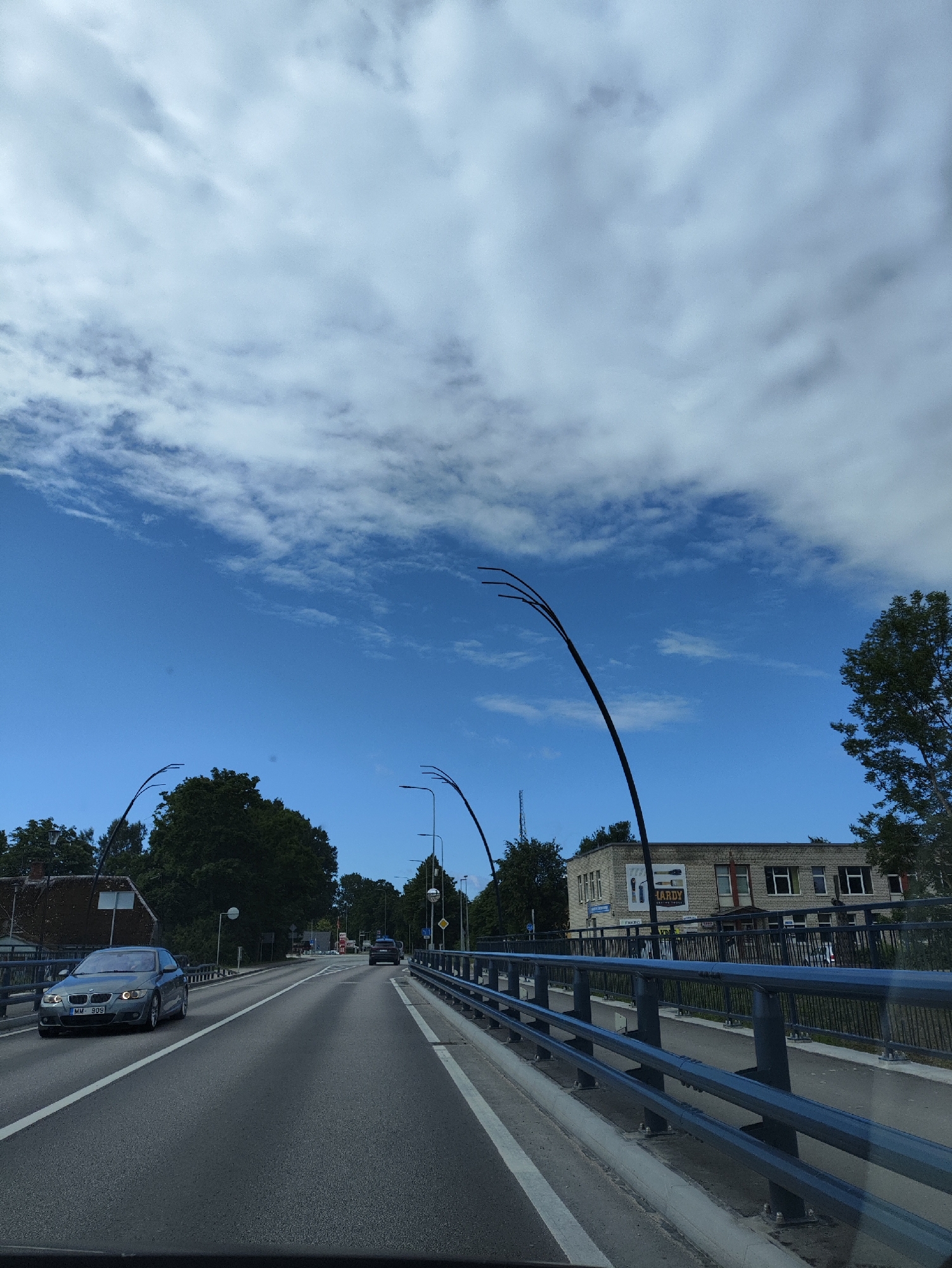 The image shows a street view in Salacgrīva, Latvia, captured from a moving vehicle. The scene features a two-lane road, with a grey BMW sedan in the foreground on the left lane, license plate MM-909. Another vehicle is visible further down the road in the right lane. Both lanes are bordered by a dark metal railing.
On the right side of the road, a two-story brick building with multiple windows is visible, with a sign that appears to read "HARDY". Trees and other buildings can be seen in the background. The sky is a bright blue with scattered white clouds. The image conveys a sense of travel through a suburban or semi-urban area on a clear day.