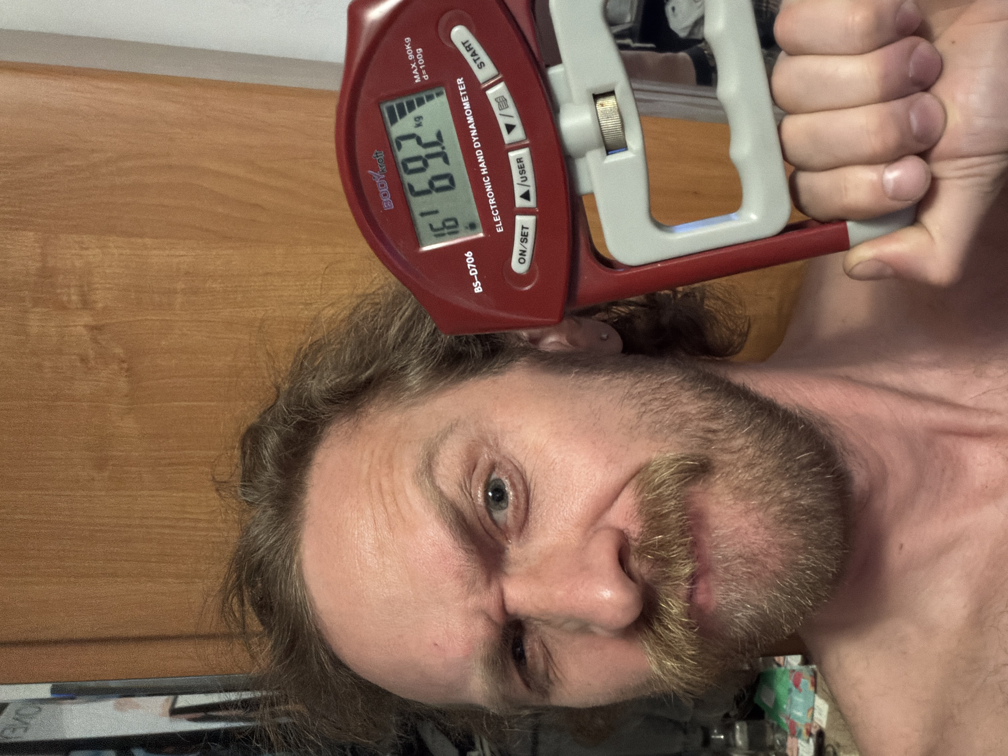 A person is shown in a selfie. The person has fair skin, a reddish-brown beard and mustache, and long brown hair. The person is holding an electronic hand dynamometer, specifically a "BODYkroft BS-D706 ELECTRONIC HAND DYNAMOMETER". The dynamometer's display reads "16 69.2 kg". The dynamometer is red and gray. The person appears to be straining while using the dynamometer. A wooden wall or door is visible in the background. A cluttered area is partially visible at the bottom left of the frame. There is no information available about the location being Saint Petersburg, Russia.