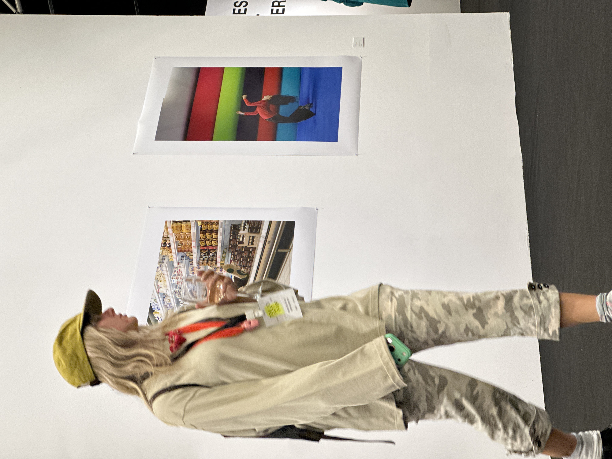 A sideways, medium indoor shot shows a woman standing in front of two posters on a white wall.
The woman faces left. She wears a yellow cap, a tan jacket, camouflage pants, and white sneakers. She holds a clear plastic container in her right hand and has a lanyard around her neck. A green phone is tucked into the jacket pocket.
Above and behind the woman are two rectangular posters attached to the wall with pushpins. The top poster is of a woman in a red shirt and black pants leaping horizontally against a background of red, green, black, cyan and blue fabric. The bottom poster shows shelves stocked with various products in a grocery store.
The background wall is white on the left and gray on the right. "ES..." and "ER..." are seen on the top of the wall.
The location is in the city of Tbilisi, Georgia.