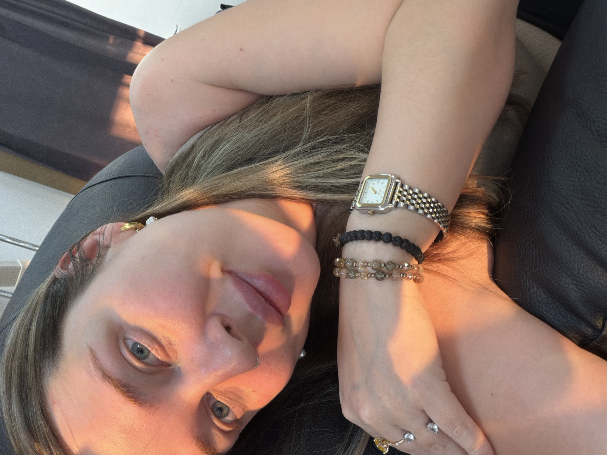 A woman with blonde hair and blue eyes is lying down, looking at the camera. She has a nose ring and earrings. On her wrist, she wears a watch, two beaded bracelets, and rings on her fingers. She is resting on a black leather surface. The lighting suggests a golden hour. The location is unknown and cannot be determined from the image.