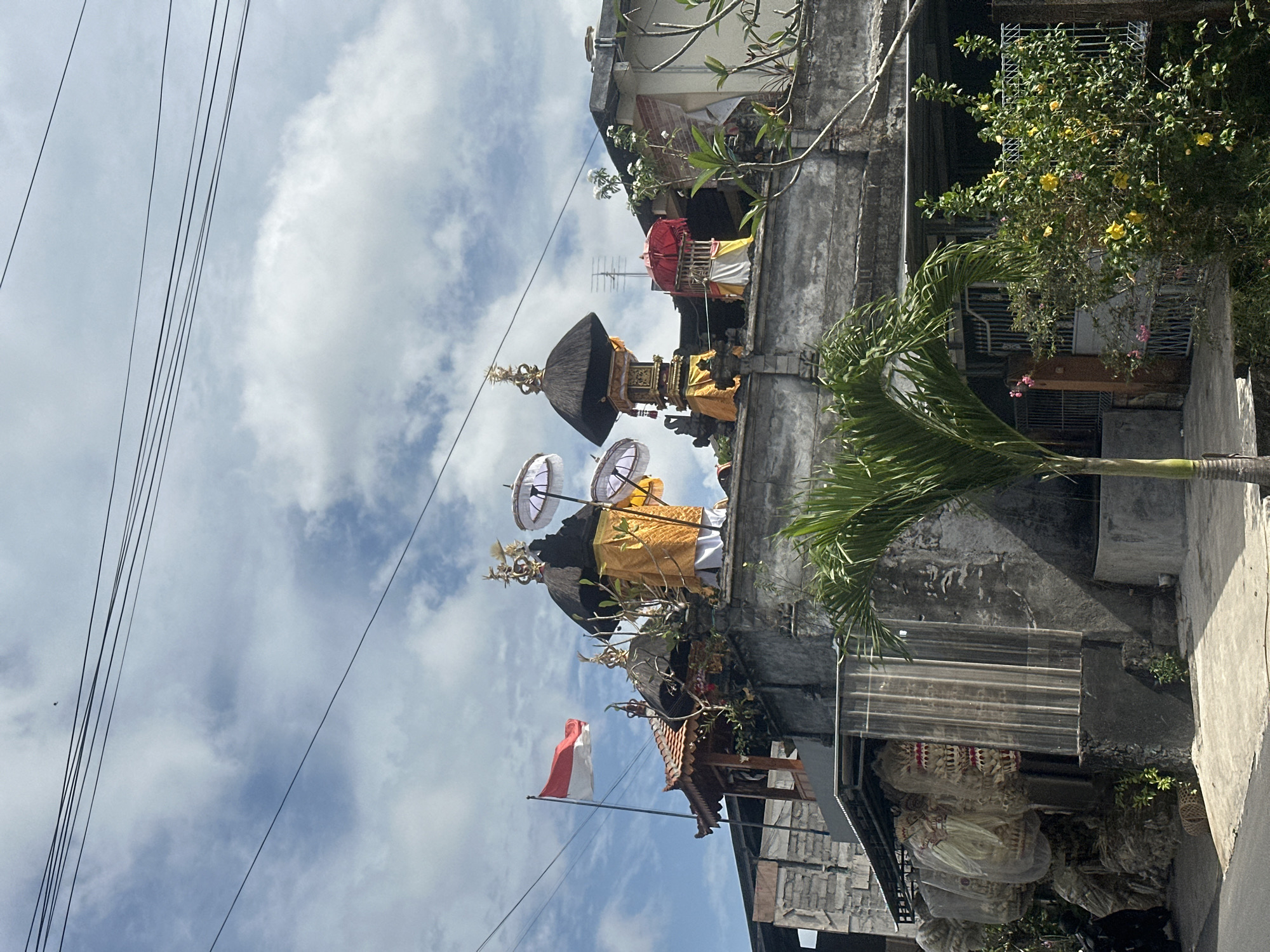 The image shows a rooftop with multiple Balinese temple structures and decorative elements, set against a cloudy sky with power lines. On the far left, a red and white Indonesian flag flies from a pole. The temple structures feature thatched roofs, gold-colored ornamentation, and yellow and white fabric coverings. Several white parasols are also visible. In the foreground, a palm tree and lush green foliage partially obscure the view of the buildings below. To the left of the temple structures, a wall displays what appear to be large, woven baskets covered in plastic. The overall scene suggests a cultural or religious site in an urban or semi-urban setting in North Kuta, Indonesia.