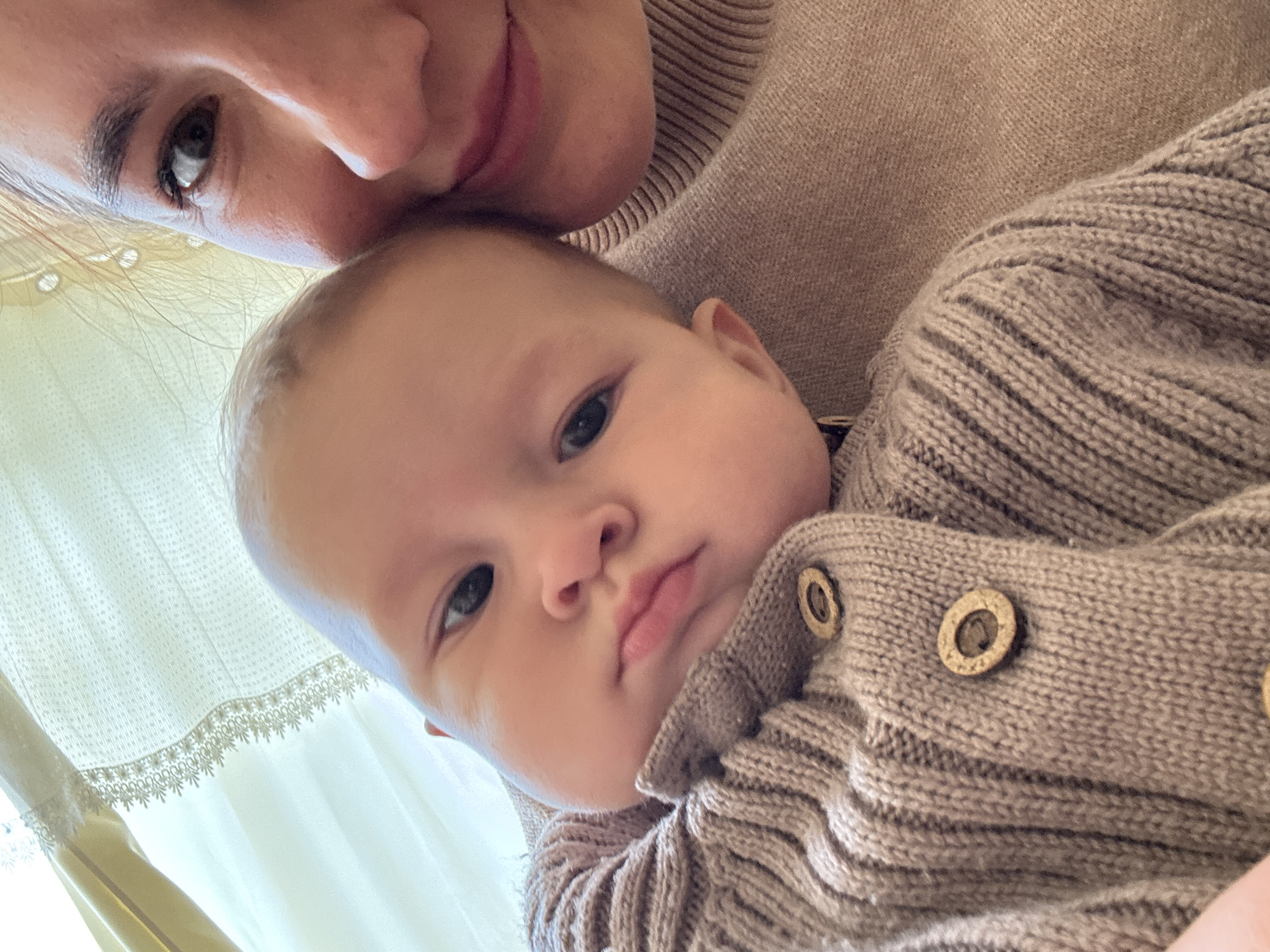 A close-up, angled shot depicts a woman and a baby. The woman's face is partially visible at the top of the frame, with her eye and mouth in focus. The baby is centered in the image, looking directly at the camera with a slightly pensive expression. Both the woman and baby are wearing similar beige-toned, knitted sweaters. The sweater worn by the baby has visible round buttons. The background is soft, with a white curtain visible in the lower left corner of the frame. The location, Sochi, Russia, cannot be verified from the visual information.