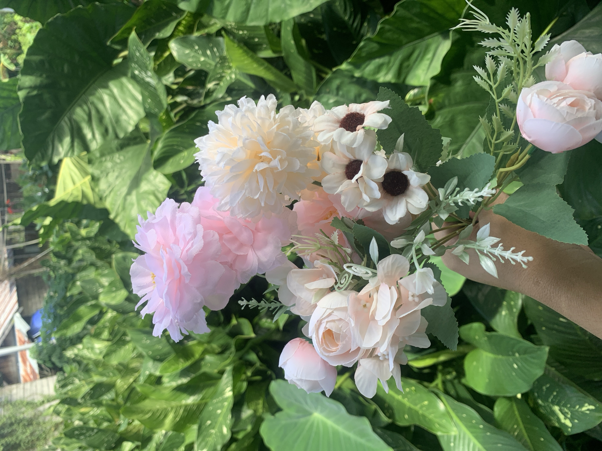 A bouquet of artificial flowers is held in a person's hand against a backdrop of lush green foliage and some out-of-focus buildings. The bouquet contains various types of flowers in shades of pink and cream, including roses, peonies, and small daisy-like flowers. The person's hand is a light brown skin tone, and their wrist is visible. The environment appears to be outdoors, possibly a garden or yard, with large leaves and some structures with corrugated metal roofs in the background. The lighting suggests it is daytime, with sunlight filtering through. The scene is static.