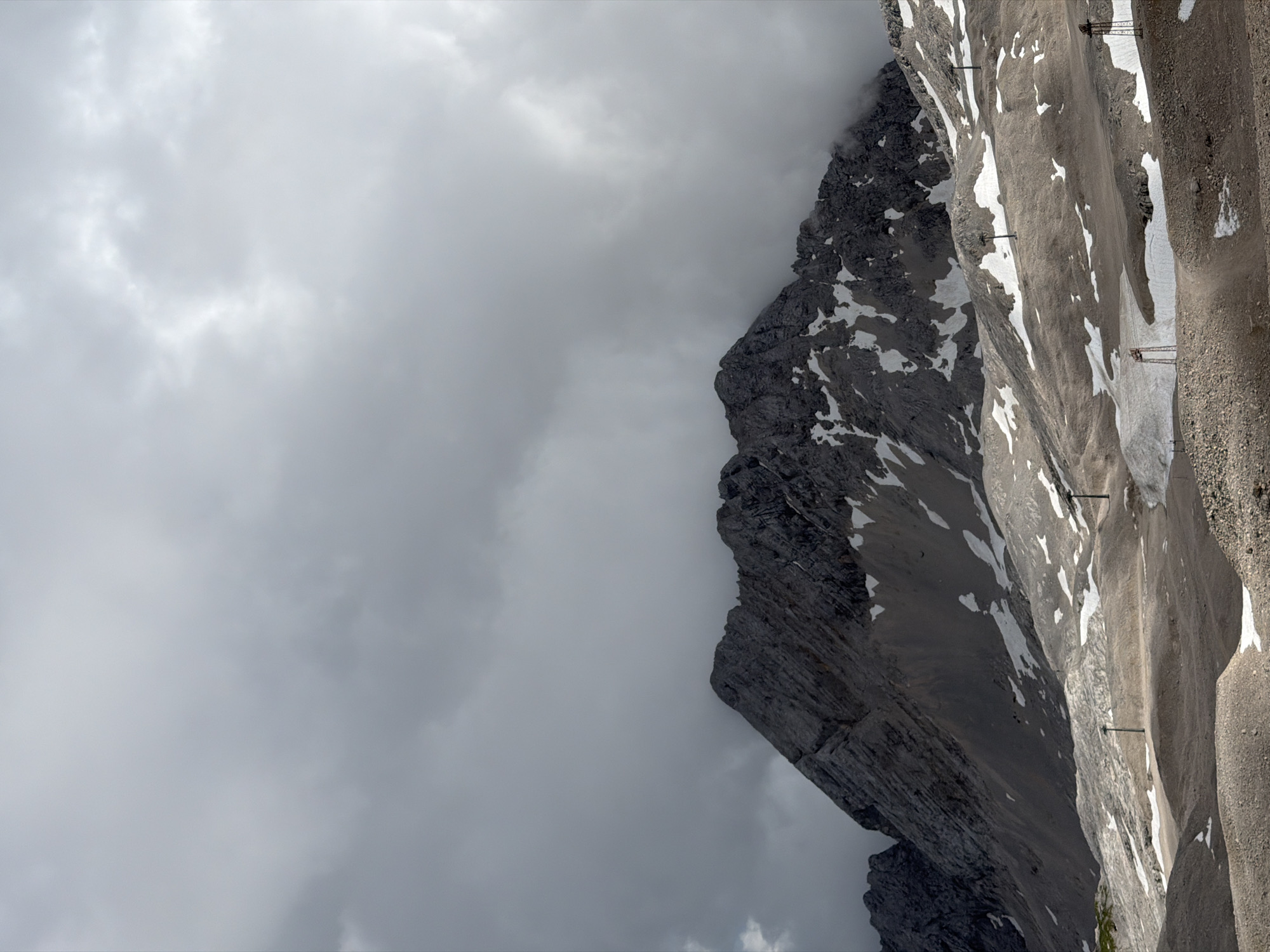 The image depicts a panoramic view of the Zugspitze massif, Germany's highest peak, located near Garmisch-Partenkirchen. The mountain is characterized by its rugged, rocky slopes, partially covered by snow and patches of ice. The lower slopes feature a barren, gravelly terrain, interspersed with snowfields. Several ski lift pylons are visible throughout the landscape, suggesting its use as a ski resort. The sky above is overcast with heavy, dark clouds, creating a dramatic and somber atmosphere. The lighting is subdued, with some areas of the mountain illuminated by shafts of light breaking through the clouds. The scene conveys a sense of rugged, alpine beauty under challenging weather conditions.