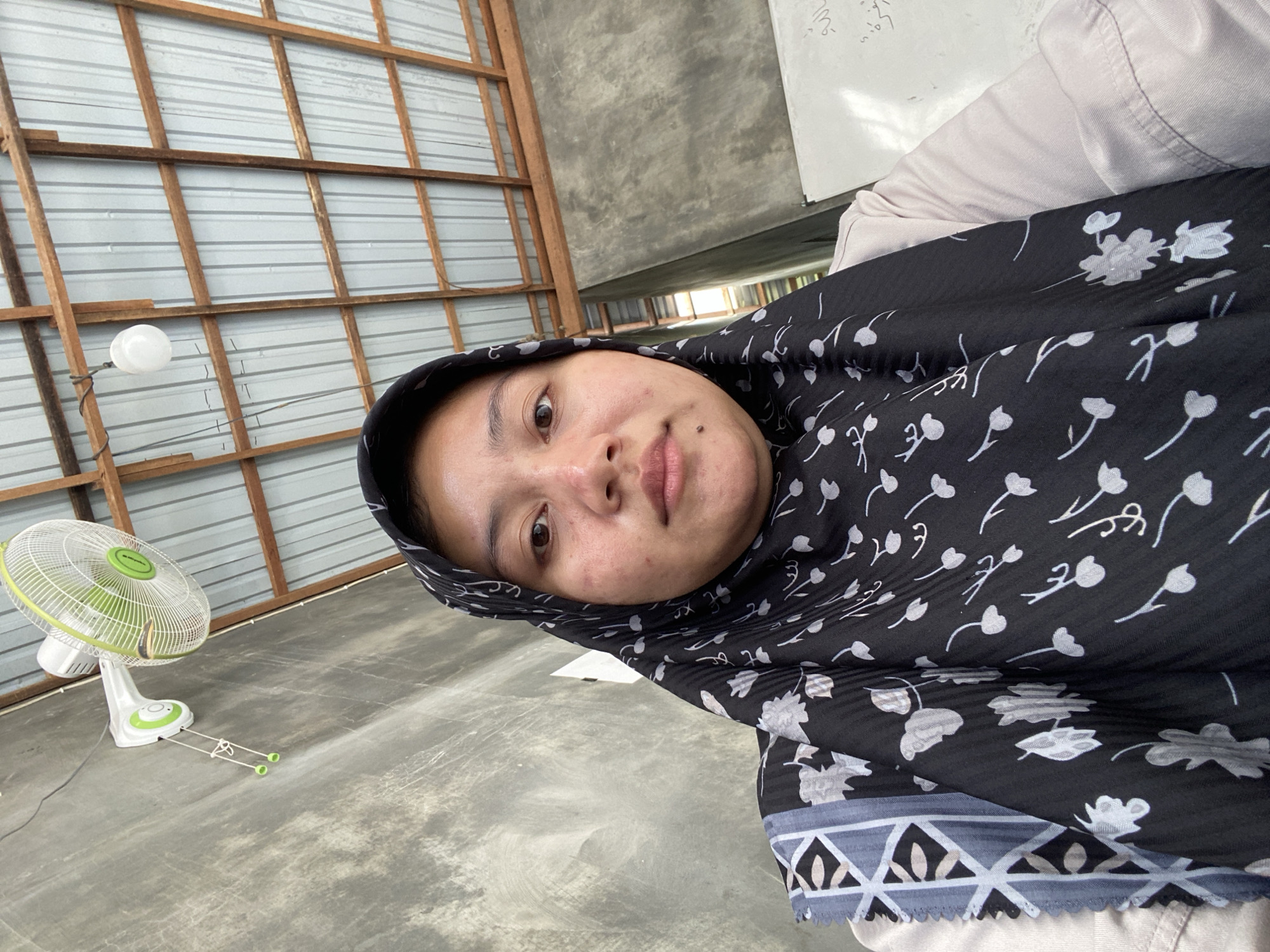 A selfie taken from a low angle shows a young woman with dark hair, wearing a black hijab with white floral patterns and a light-colored shirt. She is looking directly at the camera, with her face filling most of the frame. The background consists of a concrete wall on the left, a corrugated metal ceiling supported by wooden beams in the upper portion, and a whiteboard with Arabic writing on the right. A wall-mounted fan with green accents is visible on the left wall, and a simple light fixture hangs from the ceiling. The scene appears to be indoors, likely in a room with a rustic or unfinished aesthetic, possibly in a residential or community building. The lighting is natural, coming from an unseen source. The overall impression is that of a casual, personal photograph taken in a simple, everyday environment.