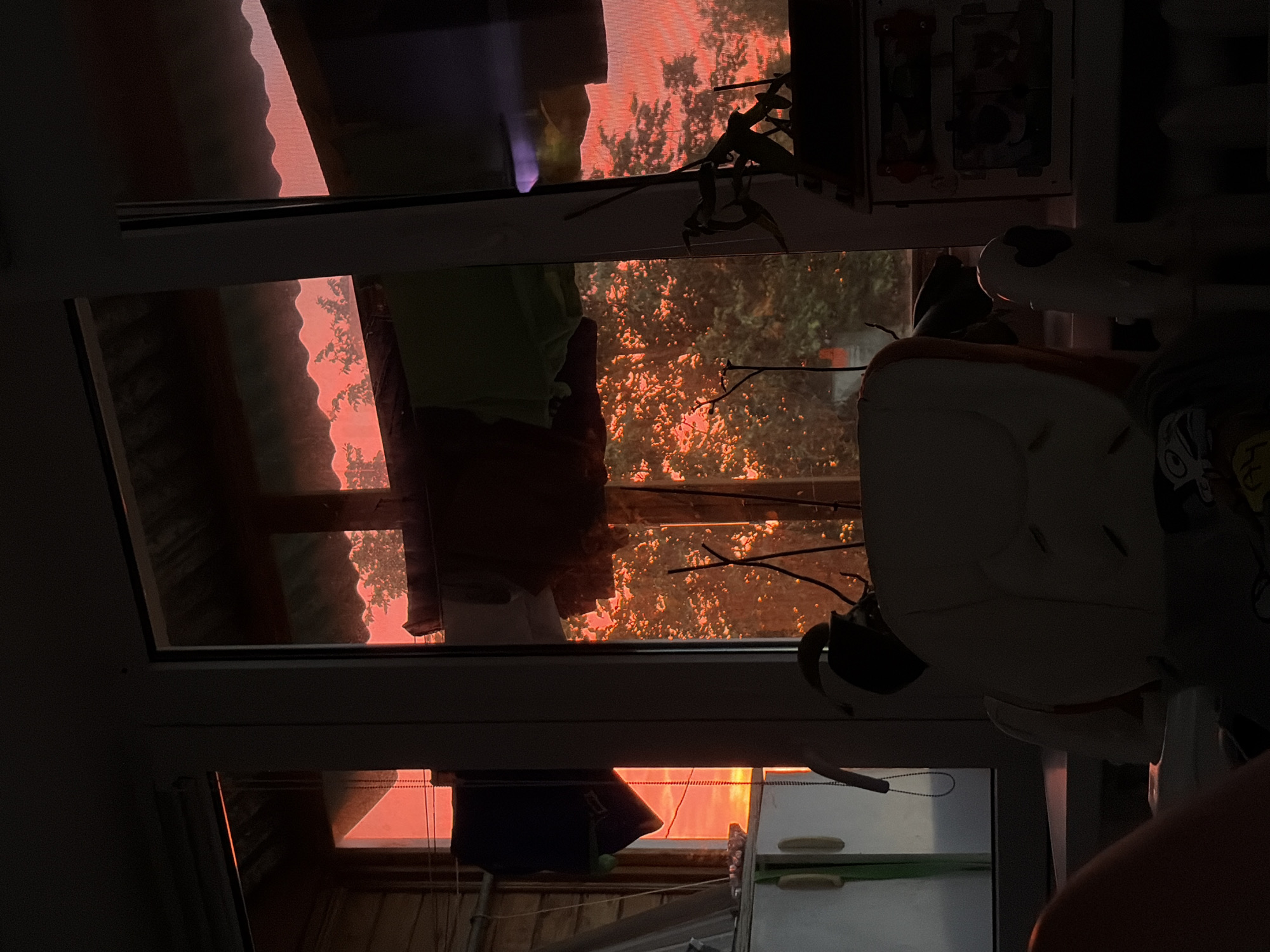 The image depicts a scene from a window in a residential building in the city of Tsentralny, Russia, during a sunset. The sky is a vibrant orange-red hue, with silhouetted trees visible in the background. Laundry is hanging on a line outside the window, suggesting a domestic setting. Inside the room, a partially visible white chair, possibly an armchair, is in the foreground. A toy or stuffed animal with white and black features is also visible. The room appears to be dimly lit, with the sunset light providing the primary illumination. The overall atmosphere is serene and somewhat melancholic, characteristic of a sunset.