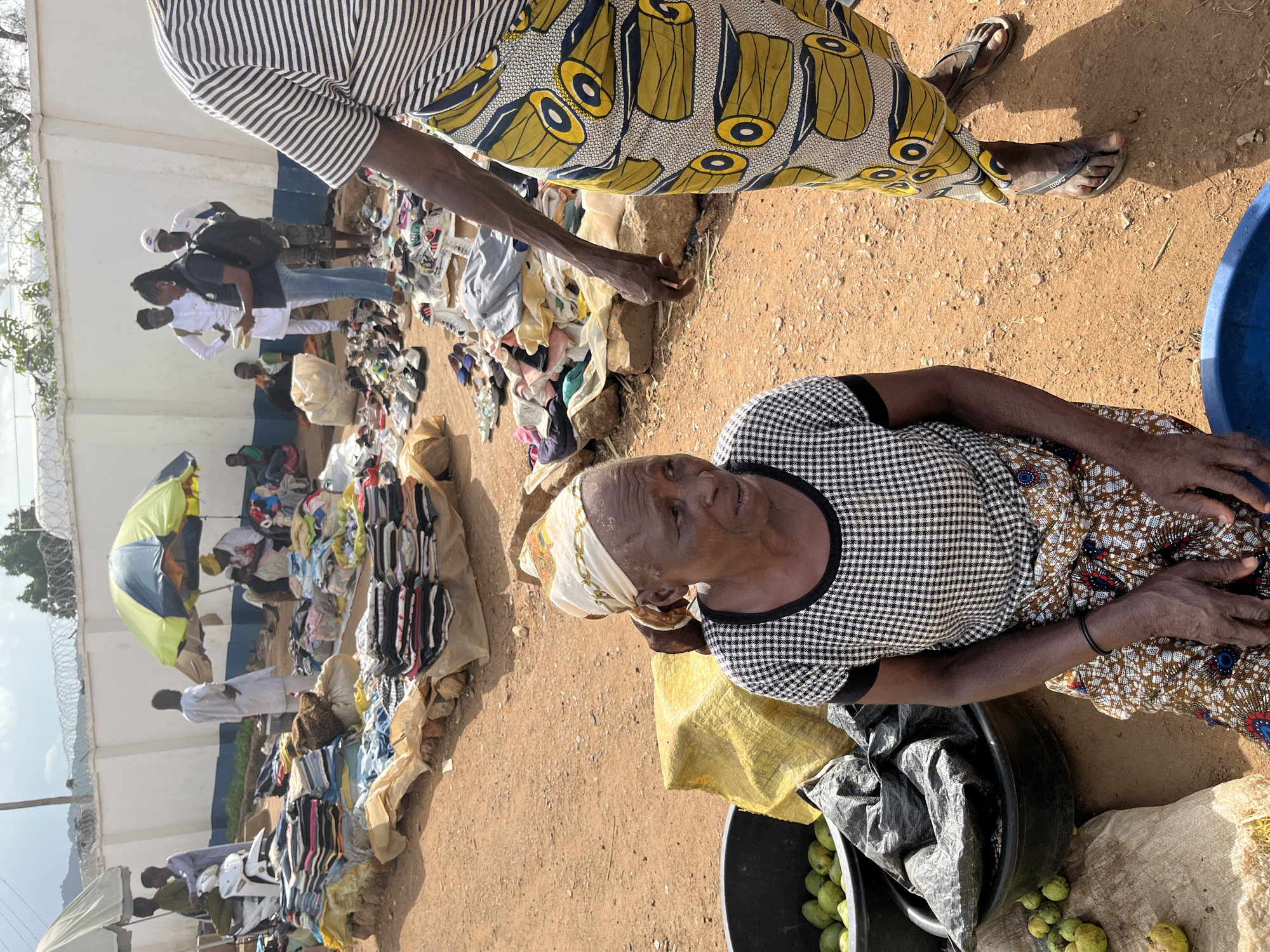 In Rafin Par, Nigeria, an outdoor market scene unfolds under a bright, likely daytime sky, judging by the strong shadows and direct sunlight. The ground is a dusty, reddish-brown earth, indicative of arid or semi-arid conditions. A white, rendered wall forms a backdrop in the distance, with a blue stripe running along its upper section. Barbed wire is visible along the top of this wall, suggesting it might be a perimeter fence for a compound or institution.
The foreground features an elderly woman wearing a black and white checkered top and a patterned skirt, with a light-colored headscarf tied around her head. She is seated next to a large black basin filled with green, unripe fruits, possibly mangoes or a similar tropical produce. Beside her, a large yellow tarp is spread out, and further to the right, a blue tub is visible, partially cut off by the frame. The woman appears to be tending to her stall, with her hands resting on her lap.
In the mid-ground and background, the market is bustling with activity. Numerous stalls are set up, displaying an array of goods, primarily clothing and footwear, neatly folded or arranged on the ground. Several people are browsing or attending to the stalls. One individual, dressed in white, is seen standing near a yellow and green umbrella, adding a splash of color to the scene. Another person, wearing a black backpack, is interacting with someone dressed in white. The overall impression is that of a vibrant local market where vendors and shoppers are engaged in commerce. The presence of various clothing items, from shoes to folded garments, suggests a diverse range of merchandise is available.