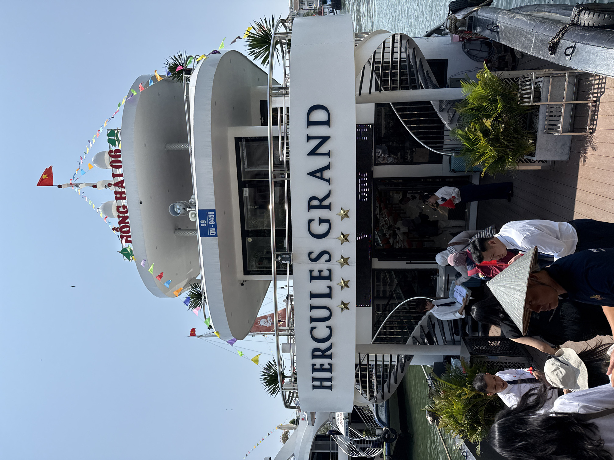 The image depicts the Hercules Grand cruise ship docked in Tuan Chau Ward, Vietnam. The ship's name, "Hercules Grand," is prominently displayed on a vertical sign with three stars underneath. The ship has multiple decks adorned with colorful triangular flags. Passengers, including some wearing traditional conical hats, are boarding the ship.