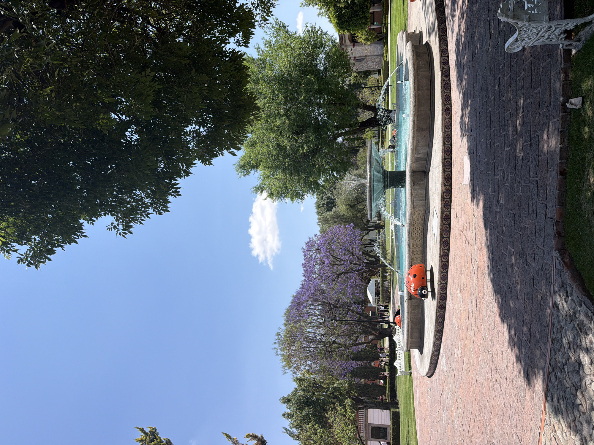 Here's a concise description of the image:
The photo shows a park setting, most likely in spring based on the blossoming trees. A prominent fountain with multiple tiers sits beside a brick path. Trees with purple blooms frame the scene. A decorative orange object resembling a ladybug is near the fountain. Buildings and people are visible in the background.