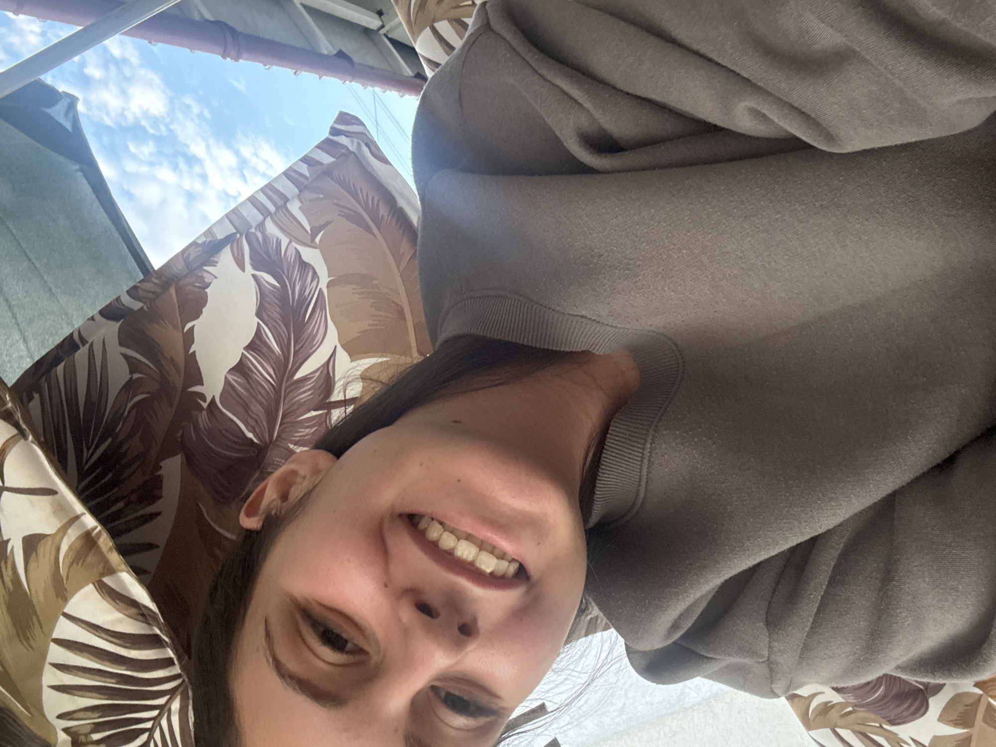 The image is a selfie taken by a young woman outdoors, likely in Bulgarovo, Bulgaria. She is leaning back, with her head tilted and a wide smile on her face, showing her teeth. The woman has dark hair and is wearing a grey sweatshirt. In the background, there is a glimpse of a blue sky with some clouds, and portions of a patterned fabric, possibly from an awning or umbrella, with large brown and cream leaves. Part of a roof structure is also visible. The overall setting suggests an informal outdoor gathering or relaxation area.