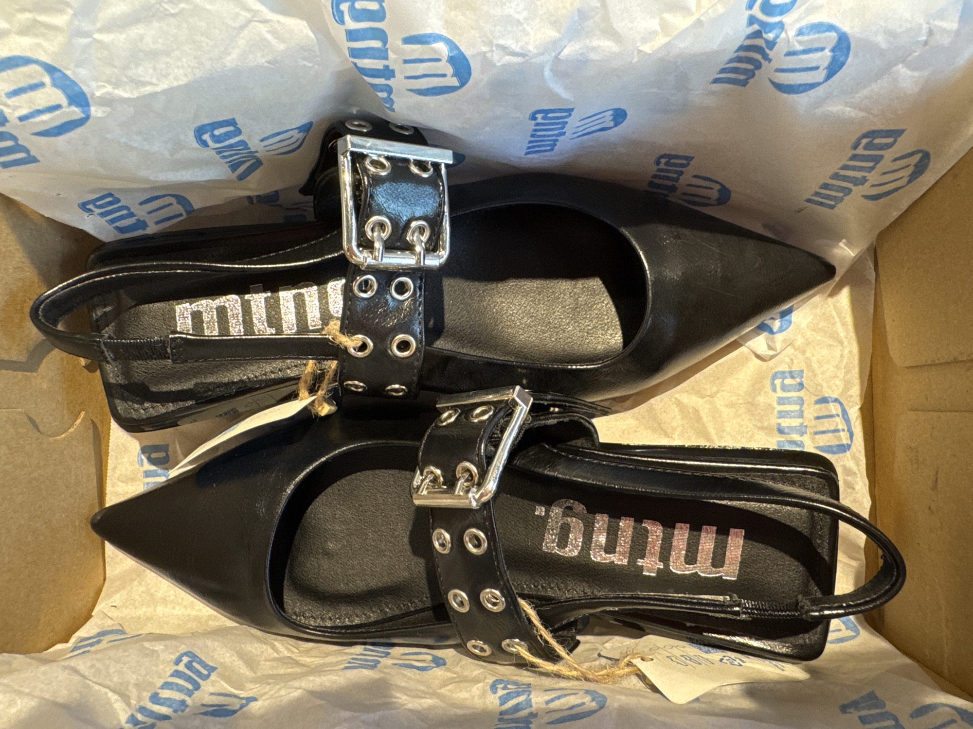 A pair of black, pointed-toe women's shoes with ankle straps and silver buckles is presented in a cardboard box lined with white tissue paper printed with a blue logo. The shoes appear new, with a small tag attached to the left shoe by twine. The logo on the tissue paper is partially visible, showing what appears to be the letters "mtnm" or "winm" repeated. The shoes are arranged side-by-side, with the right shoe positioned slightly behind and above the left shoe. The lighting suggests an indoor setting, possibly a retail environment or storage area. There are no people or animals visible in the image, nor any indication of the specific location in Aldaia, Spain.