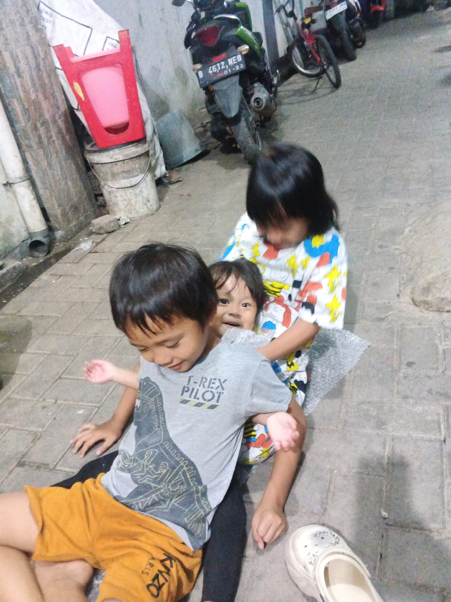 In Kadu, Indonesia, a dimly lit outdoor scene depicts three young children interacting on a paved walkway. The primary focus is on the children. The child in the foreground, a boy, is sitting with his legs extended, wearing orange shorts and a gray t-shirt with a dinosaur graphic and the words "T-REX PILOT". He is looking down with his left hand open and raised. Behind him, another child, a girl, is leaning forward, holding a younger child. The younger child, facing the camera, has a neutral expression. The girl is wearing a colorful shirt with cartoon characters. The environment includes a wall with pipes and a drain on the left, and several motorcycles parked in the background. A red plastic chair on top of a bucket is visible in the upper left quadrant. The ground is paved with square bricks.