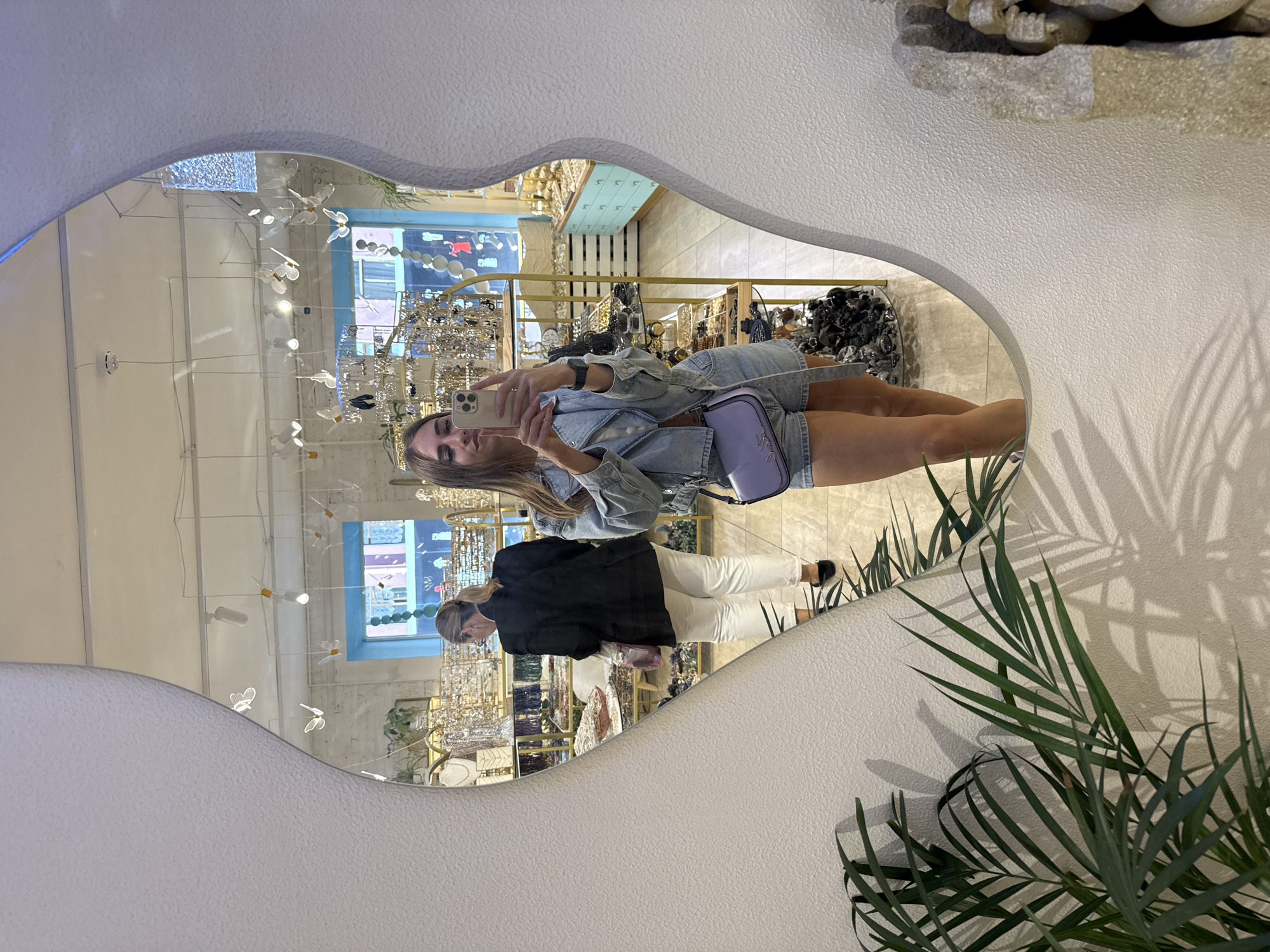 The image shows a young woman taking a selfie in a mirror within a store in Saint Petersburg, Russia. She is wearing a light blue denim jacket and shorts, with a light purple shoulder bag. Her reflection is visible in a large, irregularly shaped mirror mounted on a textured white wall. Behind her, the store's interior is reflected, featuring shelves stocked with jewelry and accessories. Another woman, wearing a black blazer and white pants, is visible in the reflection, browsing the merchandise. The store has bright lighting, with decorative butterfly-shaped lights hanging from the ceiling. A potted palm plant is visible in the foreground, casting shadows on the wall. The overall scene depicts a retail environment, likely a boutique selling jewelry and fashion accessories.