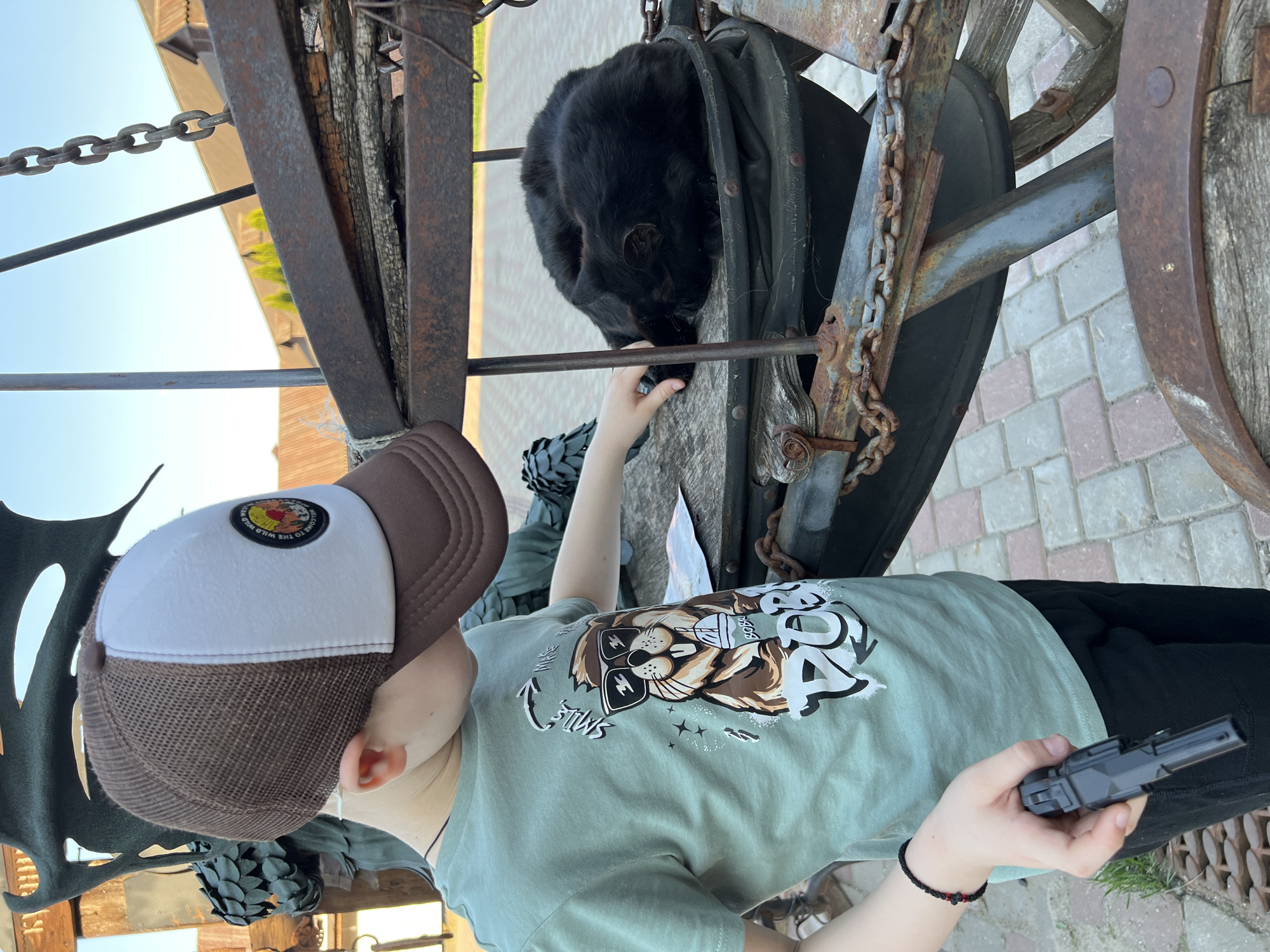A child in a brown and white baseball cap and a green T-shirt with a lion graphic is interacting with a black cat resting inside an antique carriage bellows. The child holds a small gray toy in their right hand and gently touches the cat with their left. The bellows, part of a larger carriage or cart, is situated on a paved surface that resembles the city of Sviyazhsk, Russia. A rusty chain is draped on the carriage, and other decorative ironwork and structural elements are visible.