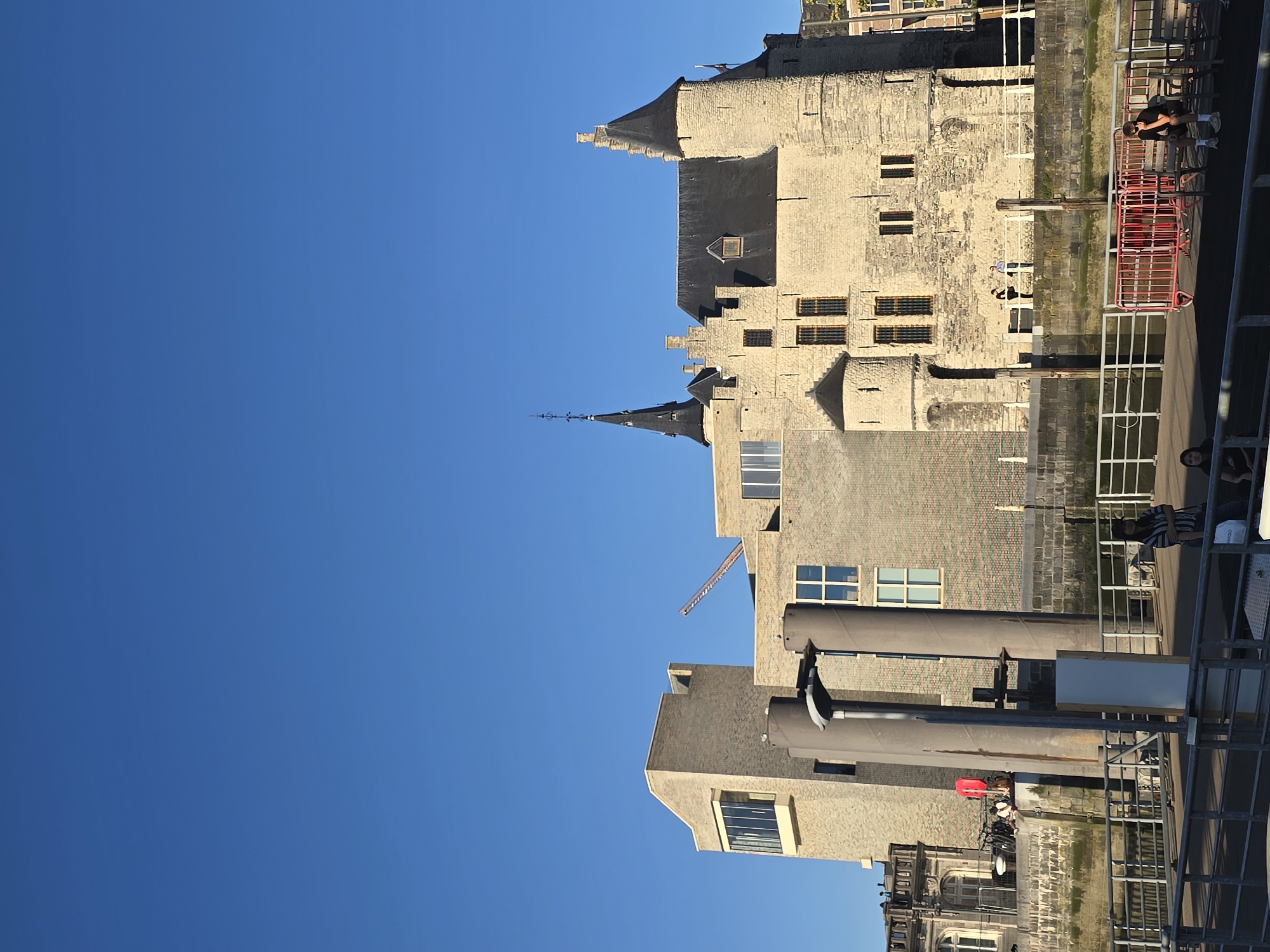 The image depicts the Het Steen castle in Antwerp, Belgium, on a clear, sunny day. The historic stone structure, with its distinctive towers and spires, is complemented by a modern brick building with large windows, likely an extension or adjacent facility. A construction crane is visible in the background, suggesting ongoing development or renovation in the area.
In the foreground, a waterfront promenade is present, bordered by a railing. Several individuals are visible on the promenade and near the castle. Some are standing and conversing, while others are seated on benches. One person is sitting on a bench in the foreground, looking down. A red metal barrier is also present along the waterfront. The Scheldt river or a canal likely flows in front of the castle, with a stone embankment visible. The overall scene conveys a sense of historical significance blended with contemporary urban life.