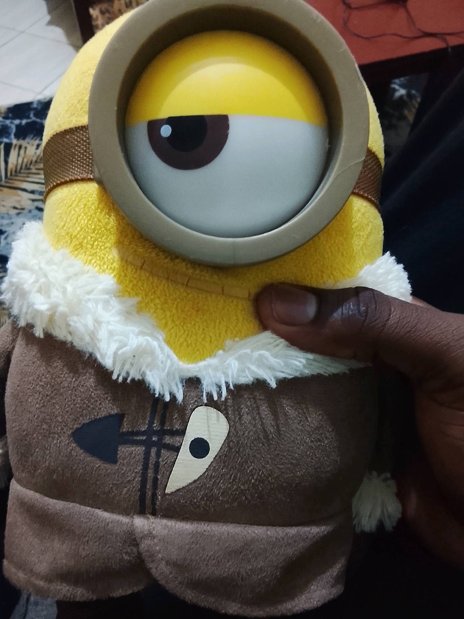 A plush toy of a Minion character from the "Despicable Me" franchise is held by a person in Ongata Rongai ward, Kenya. The toy is primarily yellow, with a large single eye covered by a goggles-like frame. It is wearing a brown fuzzy jacket with white trim. The toy is being held by a hand with dark skin. The background is blurred, but appears to be an indoor setting with some furniture and fabric visible. The image is lit by natural light.