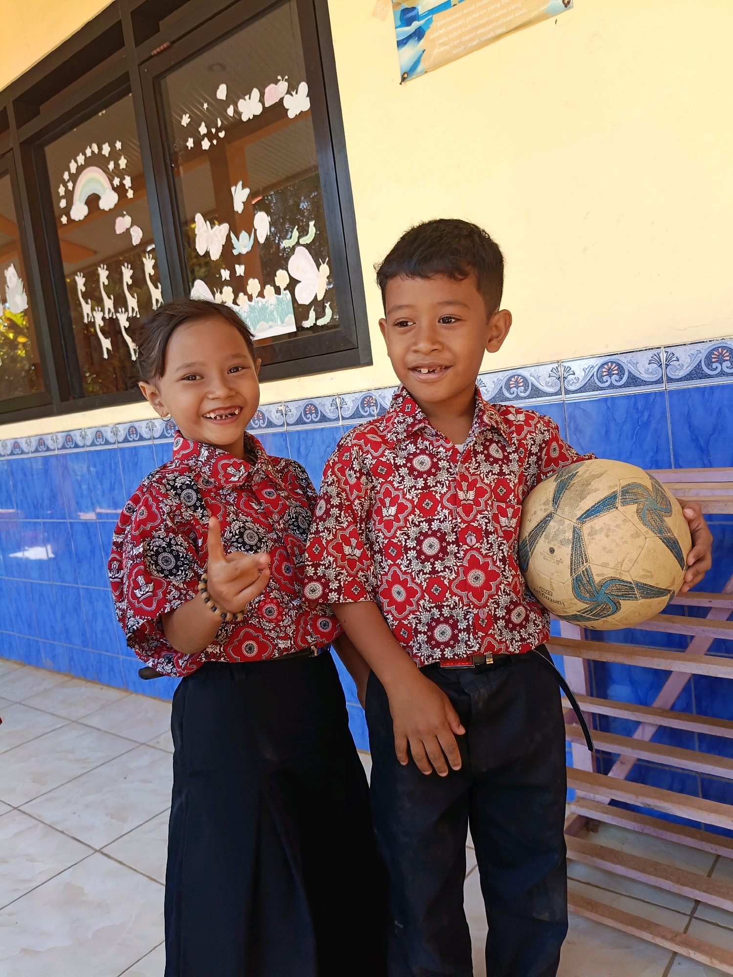 A young girl and boy, likely students at a primary school in Gunungwungkal, Indonesia, are posing for a photo. The girl, on the left, is smiling and holding up her right hand with her index and pinky fingers extended. She wears a red and white patterned shirt and a black skirt. The boy, on the right, is also smiling and holding a worn soccer ball. He is dressed in a matching red and white patterned shirt and black pants. Behind them, a building wall with blue tiled accents is visible. The wall is adorned with colorful drawings of butterflies, giraffes, and a rainbow, suggesting a school environment. A wooden bench or railing is situated to the right of the children. The image captures a candid moment, highlighting the children's youthful energy and the vibrant atmosphere of their school.